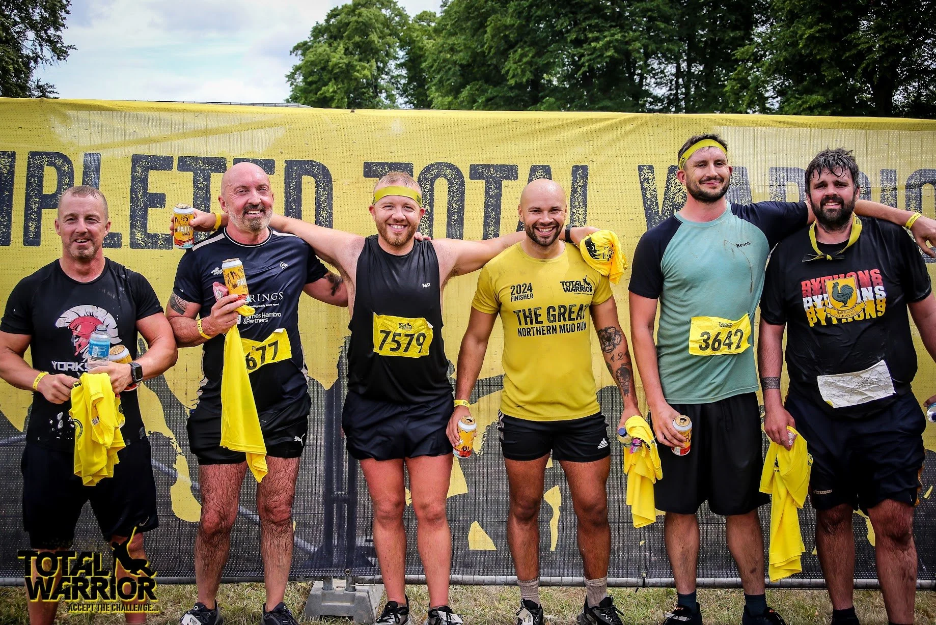 Group of six men standing together after finishing a mud run race, smiling with arms around each other, holding drinks, and wearing athletic clothes and race bibs. They are outdoors with a yellow banner in the background that says 'Total Warrior' and 'Accept the challenge.'