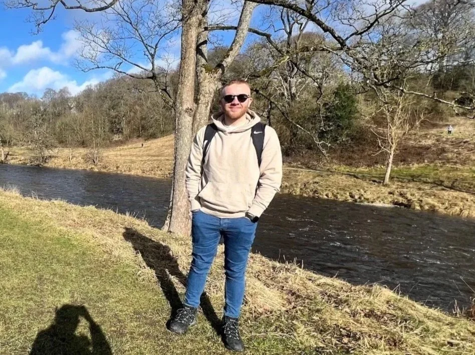 A man in sunglasses wearing a beige hoodie and blue jeans stands outdoors near a river, with trees and a grassy area in the background.