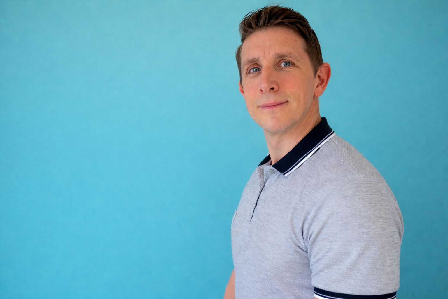 A man with short brown hair and blue eyes standing against a light blue background, wearing a light gray polo shirt with a navy collar and white stripes.