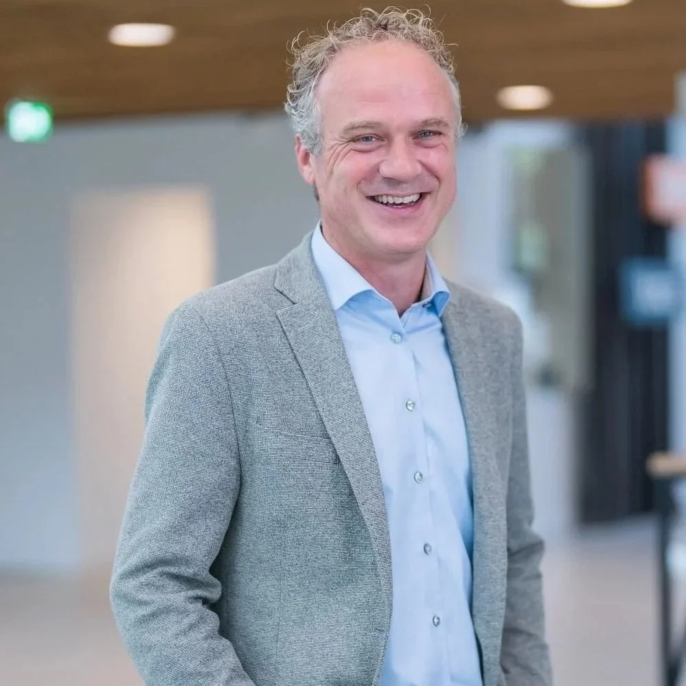 Een lachende man met kort, grijs, gekruld haar in een kantoorruimte.