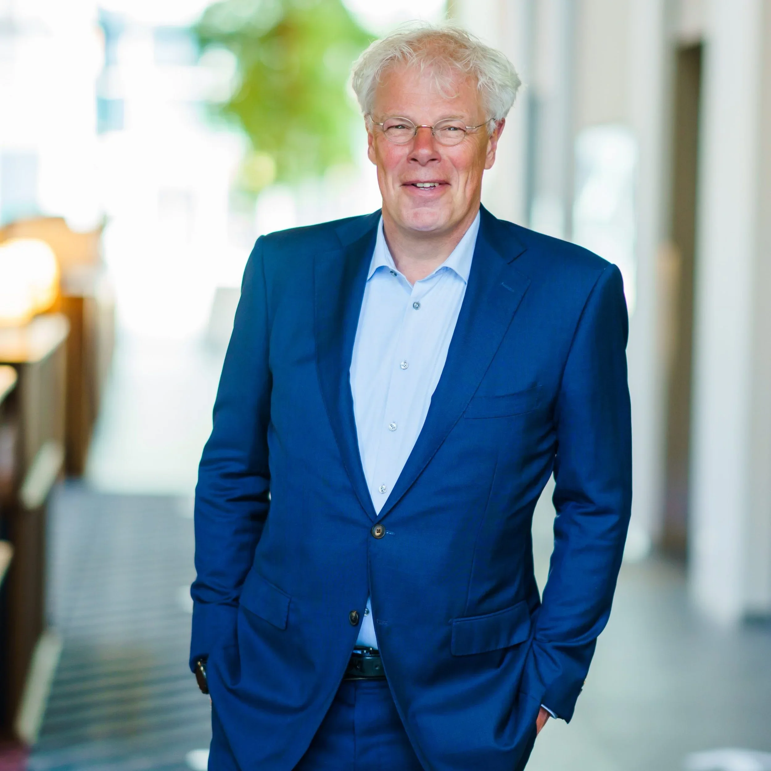 Man in een blauwe pak met lichte blauwe overhemd, glimlachend, binnen in een gebouw, met een achtergrond van planten en ramen.