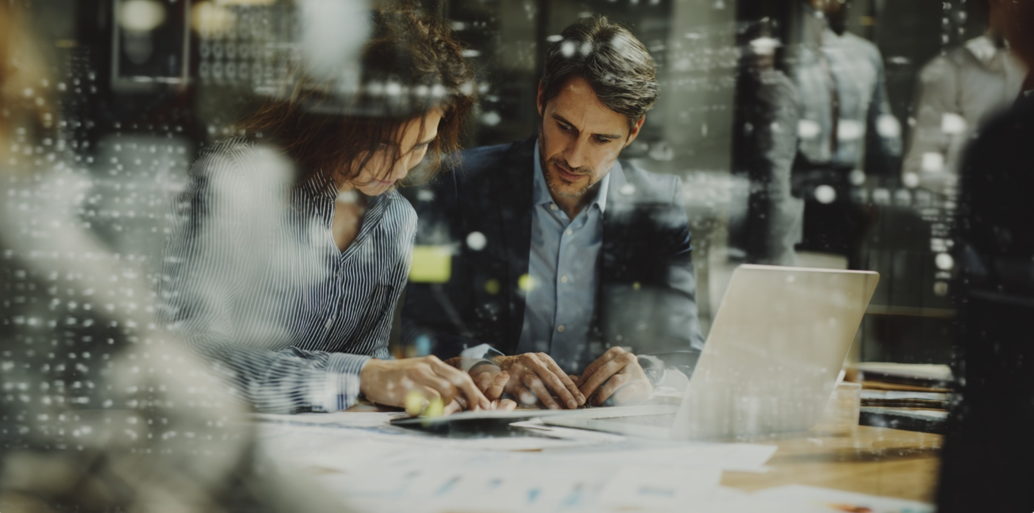 Twee zakenmensen bekijken documenten op een tafel met een laptop in een kantoor, gezien door een raam.