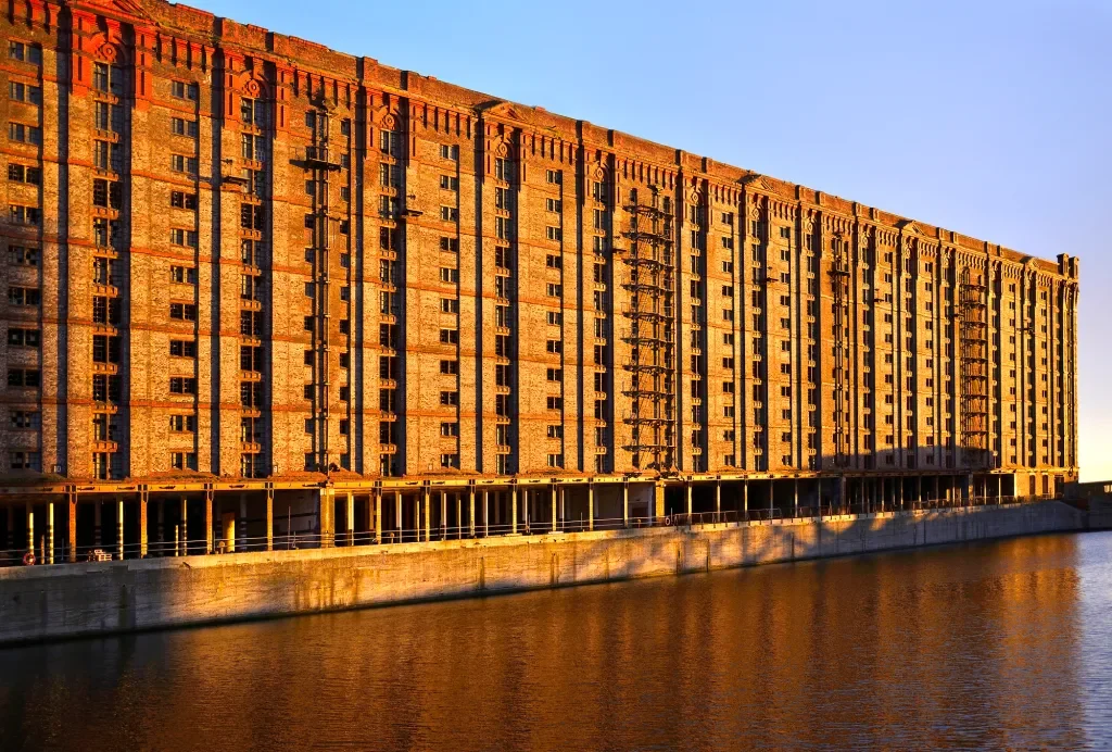 A large brick residential building with multiple floors, situated by a water canal, illuminated by warm sunlight at sunset.