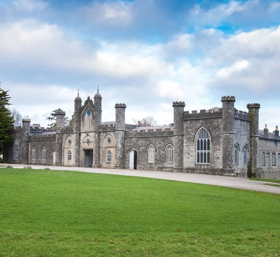 A large stone castle with battlements, turrets, and arched windows, set against a blue sky with scattered clouds, surrounded by a green lawn.