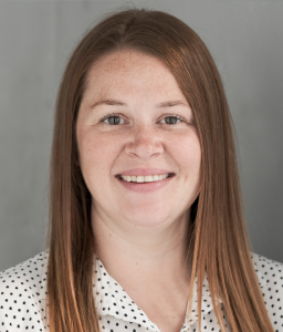 Headshot of a woman with long red hair, smiling, wearing a white blouse with black polka dots.
