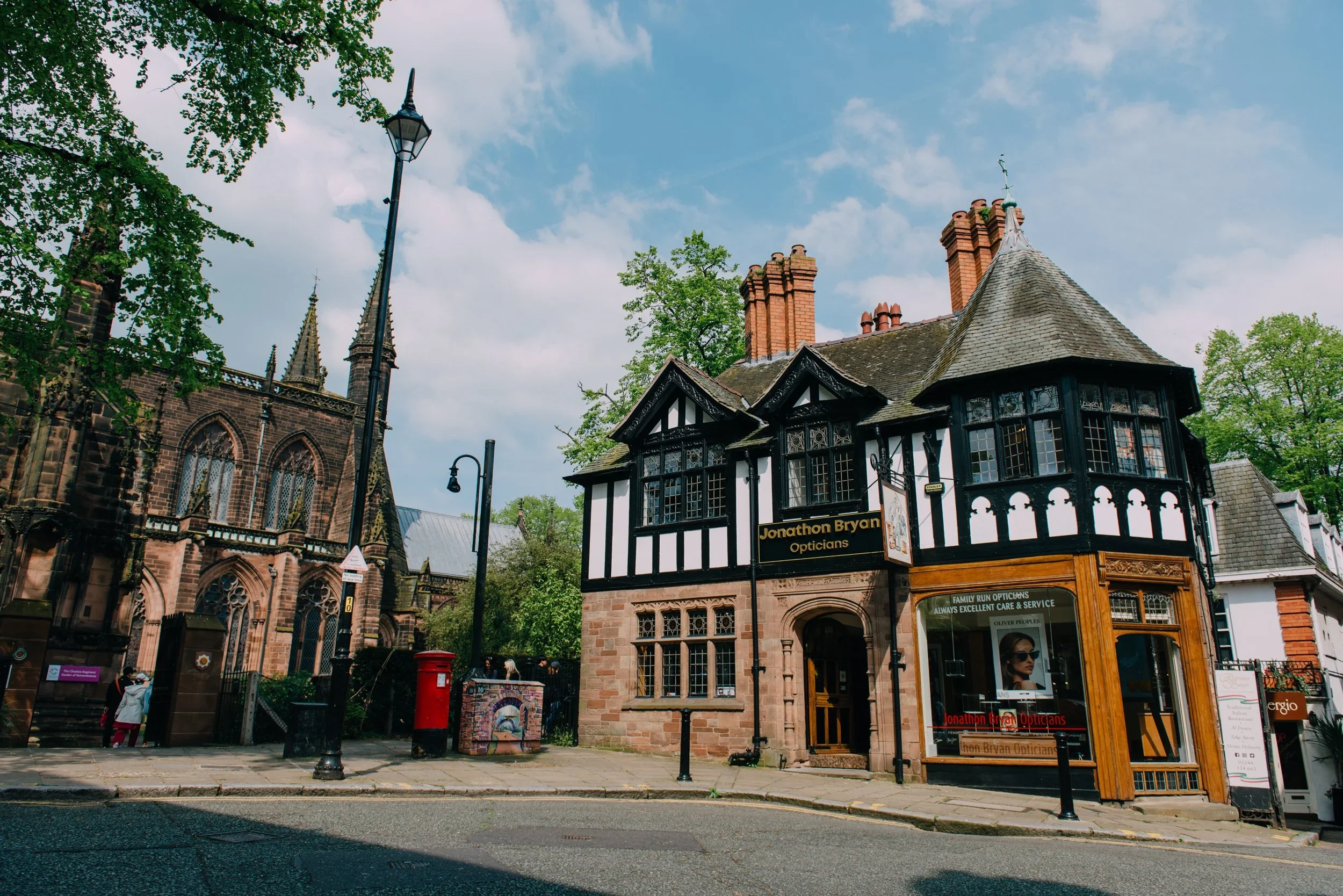 A street scene with a black and white Tudor-style optician shop and a Gothic church in the background, surrounded by green trees and a blue sky with clouds.