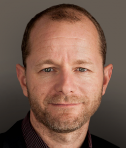 Close-up portrait of a middle-aged man with light skin, short light brown hair, and a beard, wearing a collared shirt.