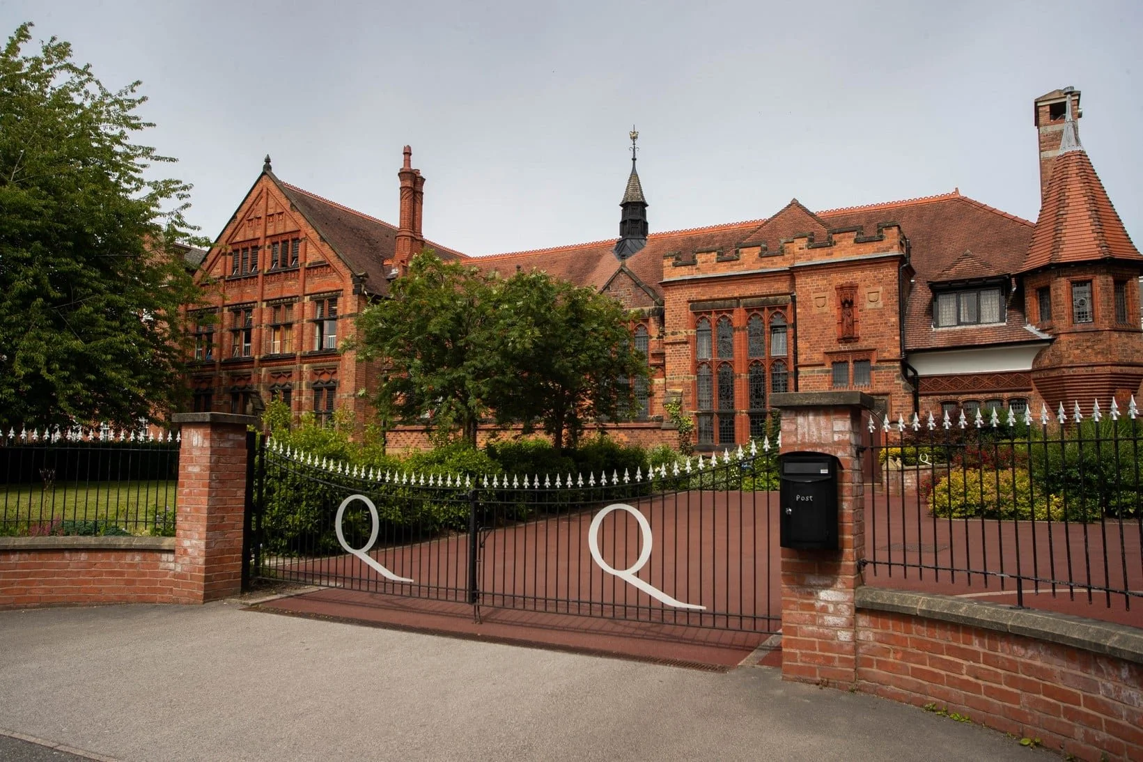 Large red brick mansion with gothic architectural elements, surrounded by a black iron fence with white decorative accents and a brick wall, green trees, a driveway, and a mailbox.