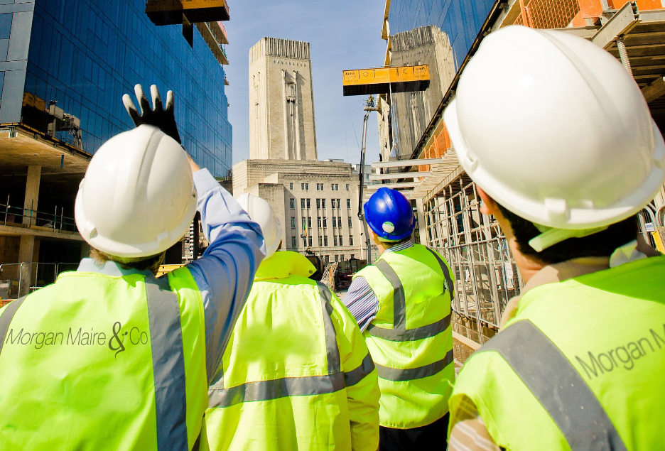 Construction workers wearing safety gear and high-visibility vests at a city construction site, with a tall building in the background.
