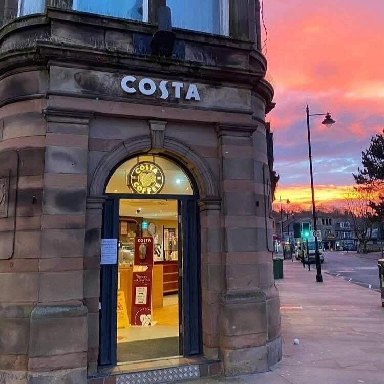 Exterior of a Costa Coffee shop on a street corner at sunset, with a decorative stone building facade, showing the open door, illuminated inside, and a sunset sky in the background.