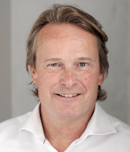 A middle-aged man with light brown hair and blue eyes, wearing a white dress shirt, smiling against a plain gray background.