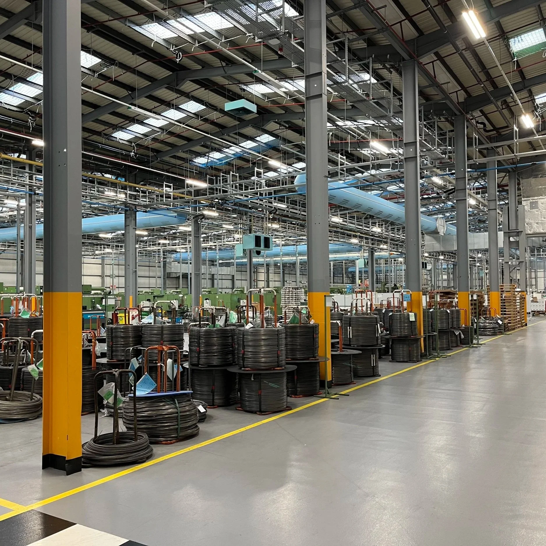 View of an industrial warehouse with shelves of rolls of black cable or wire stacked on carts, yellow safety markings on the concrete floor, and a high ceiling with metal pipes and ventilation ducts.