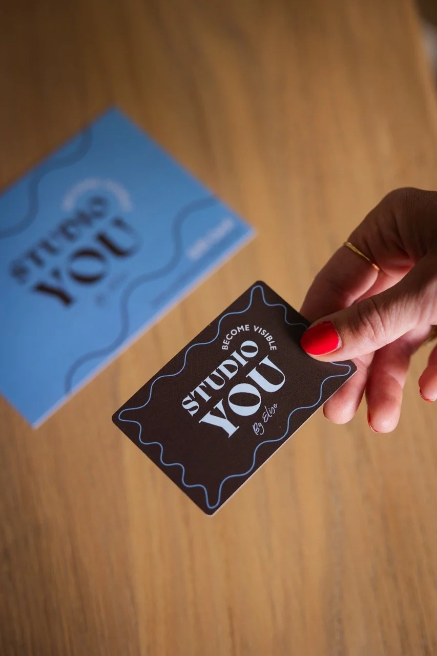 Een hand die een zwarte visitekaart vasthoudt met de tekst 'Studio You. By Elise' en een blauwe visitekaart met dezelfde tekst op de achtergrond.