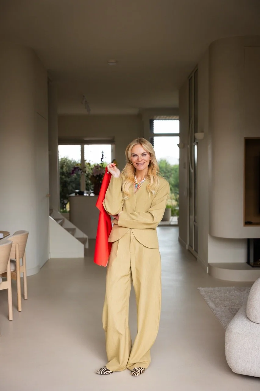 Elise draagt een beige broekpak en houdt een gekleurde doek in haar hand, binnen in een modern ingerichte studio.