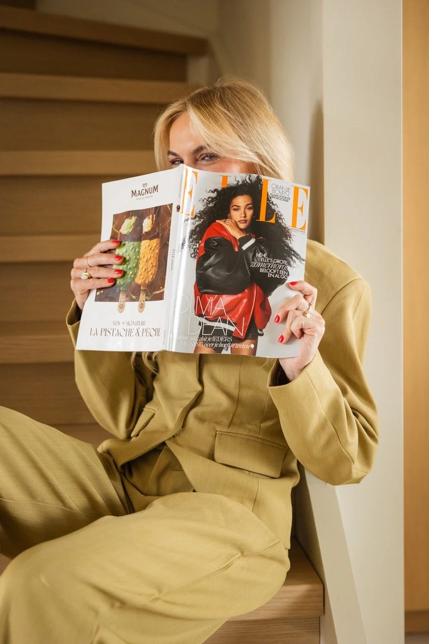 Elise in een beige pak zit op de trap en leest een Elle magazine, haar gezicht wordt deels bedekt door het magazine.
