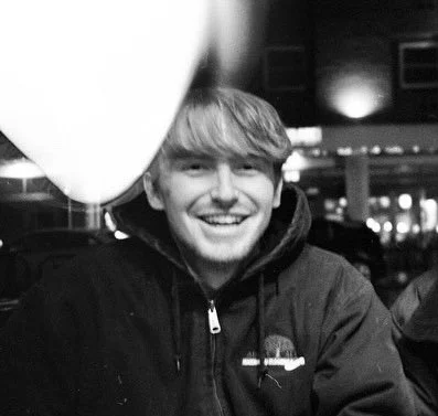A smiling young man wearing a dark jacket outdoors at night, with indistinct lights and structures in the background.