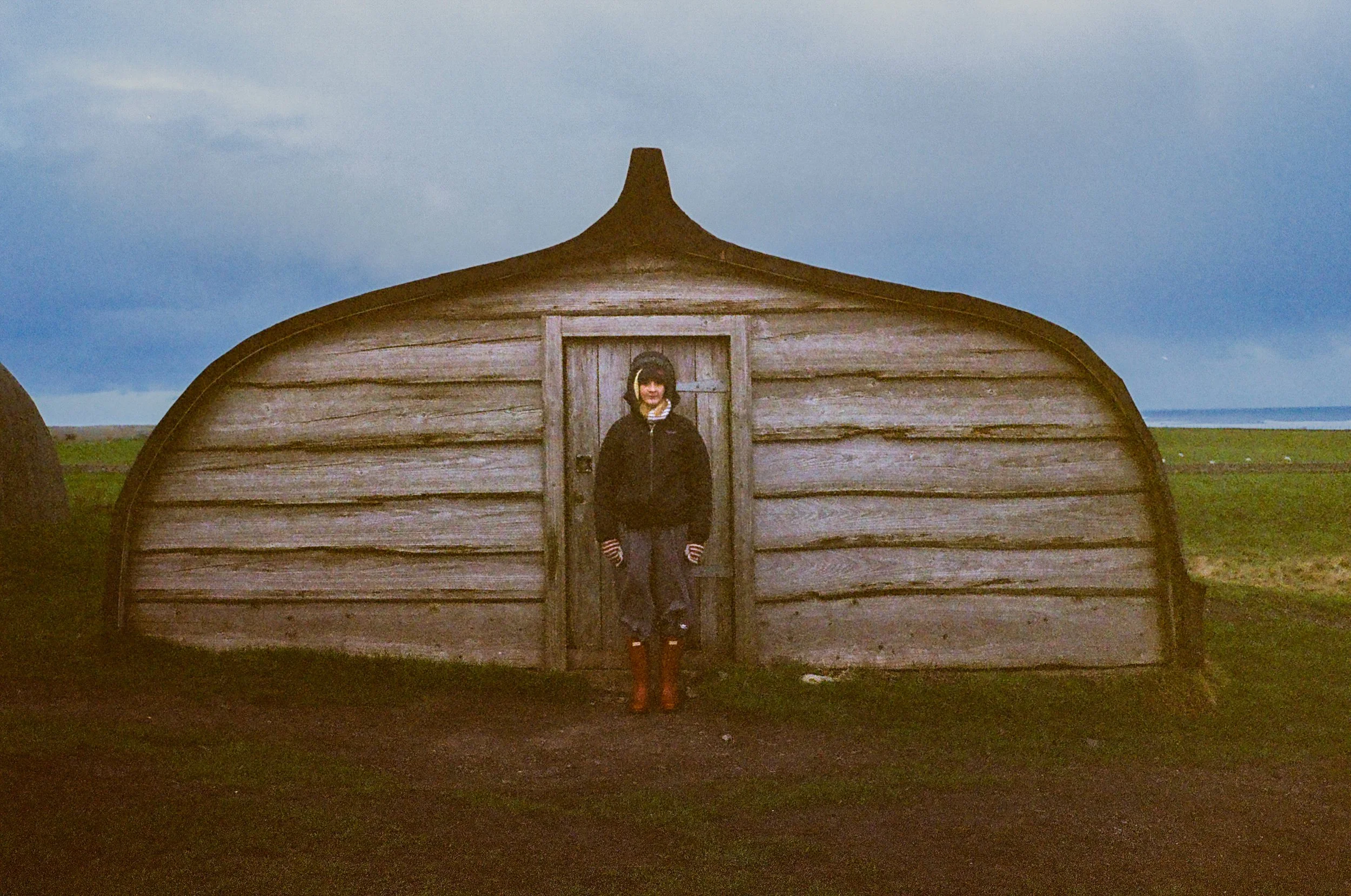 A person wearing a black jacket, striped gloves, and red boots standing in front of a small wooden house shaped like a whale.