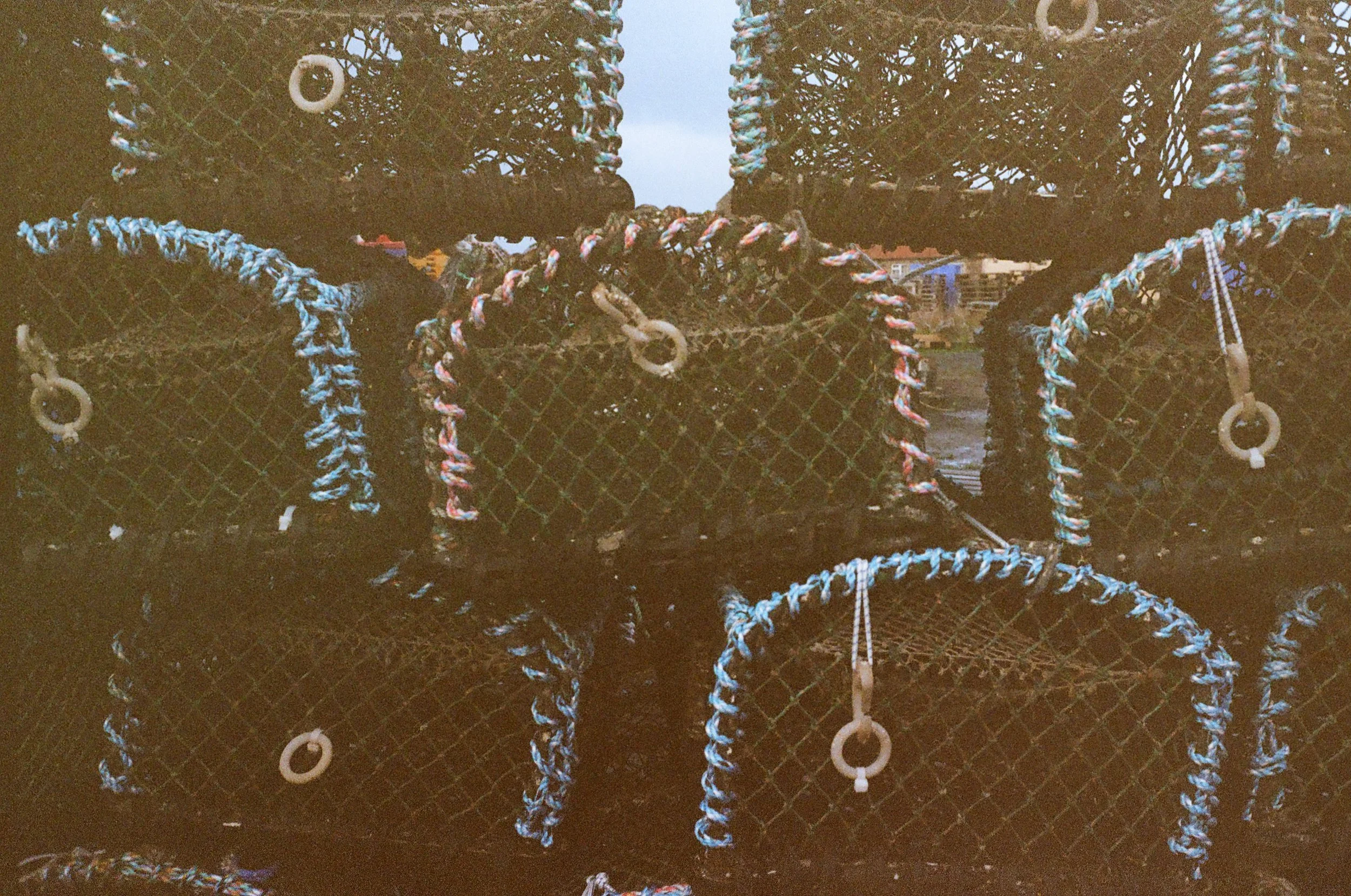 Close-up of black metal mesh containers with colorful ropes and eyelets, stacked outdoors with a cloudy sky and buildings in the background.