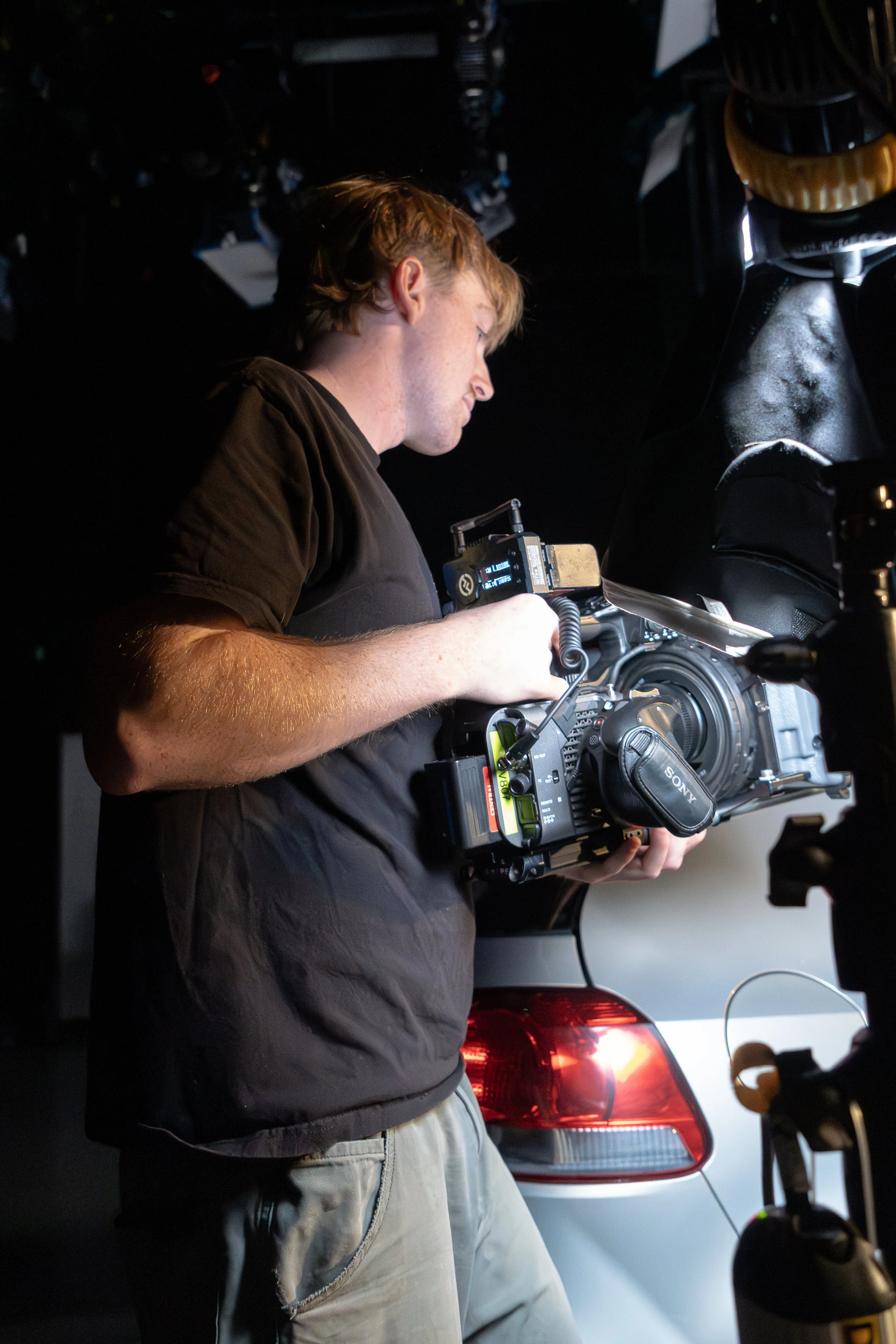 A young man operating a professional video camera inside a vehicle, with a black background and the rear of a white car visible.