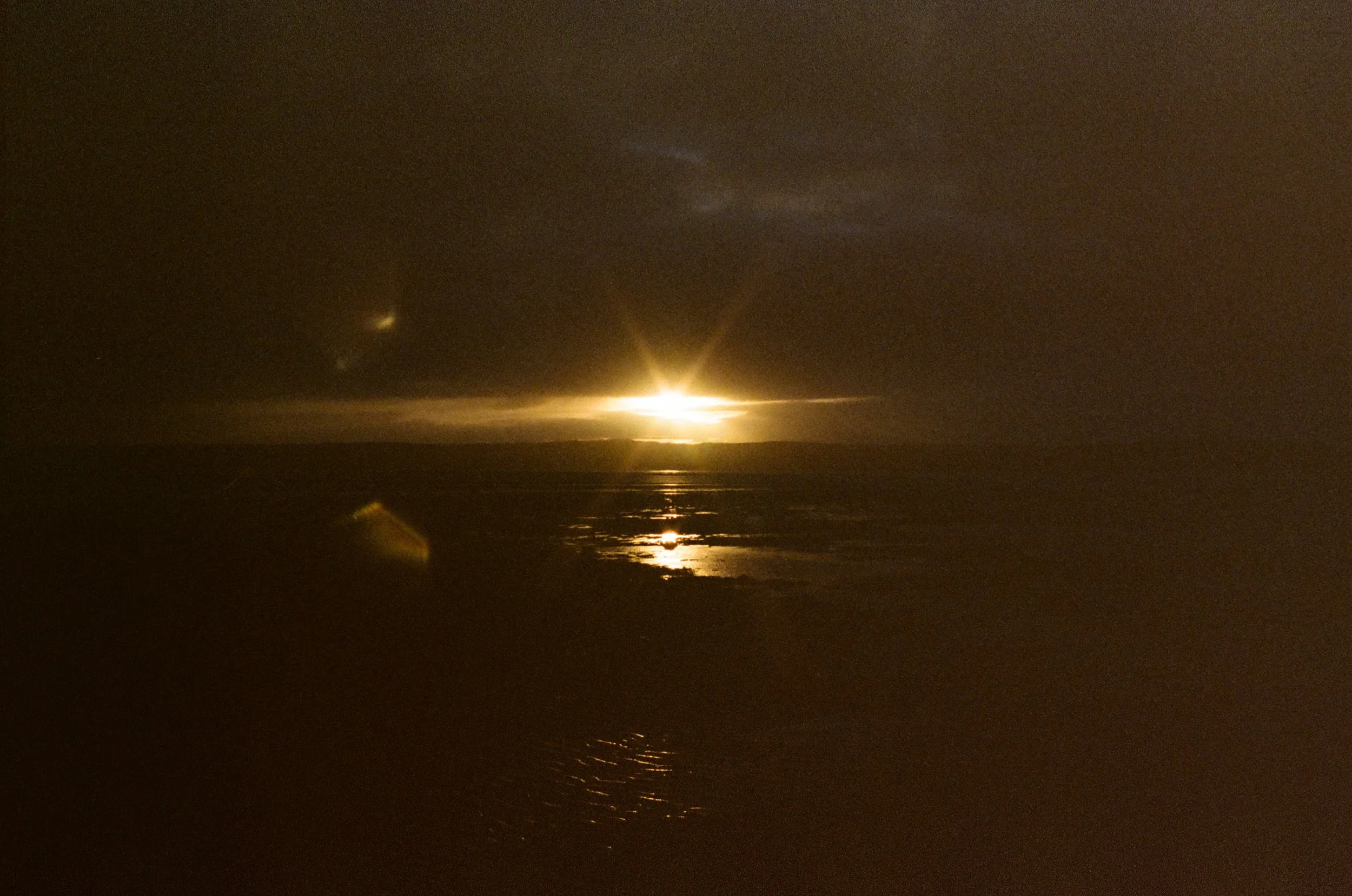 A sunset over a calm body of water, with the sun partially obscured by clouds, creating a reflection on the water's surface.