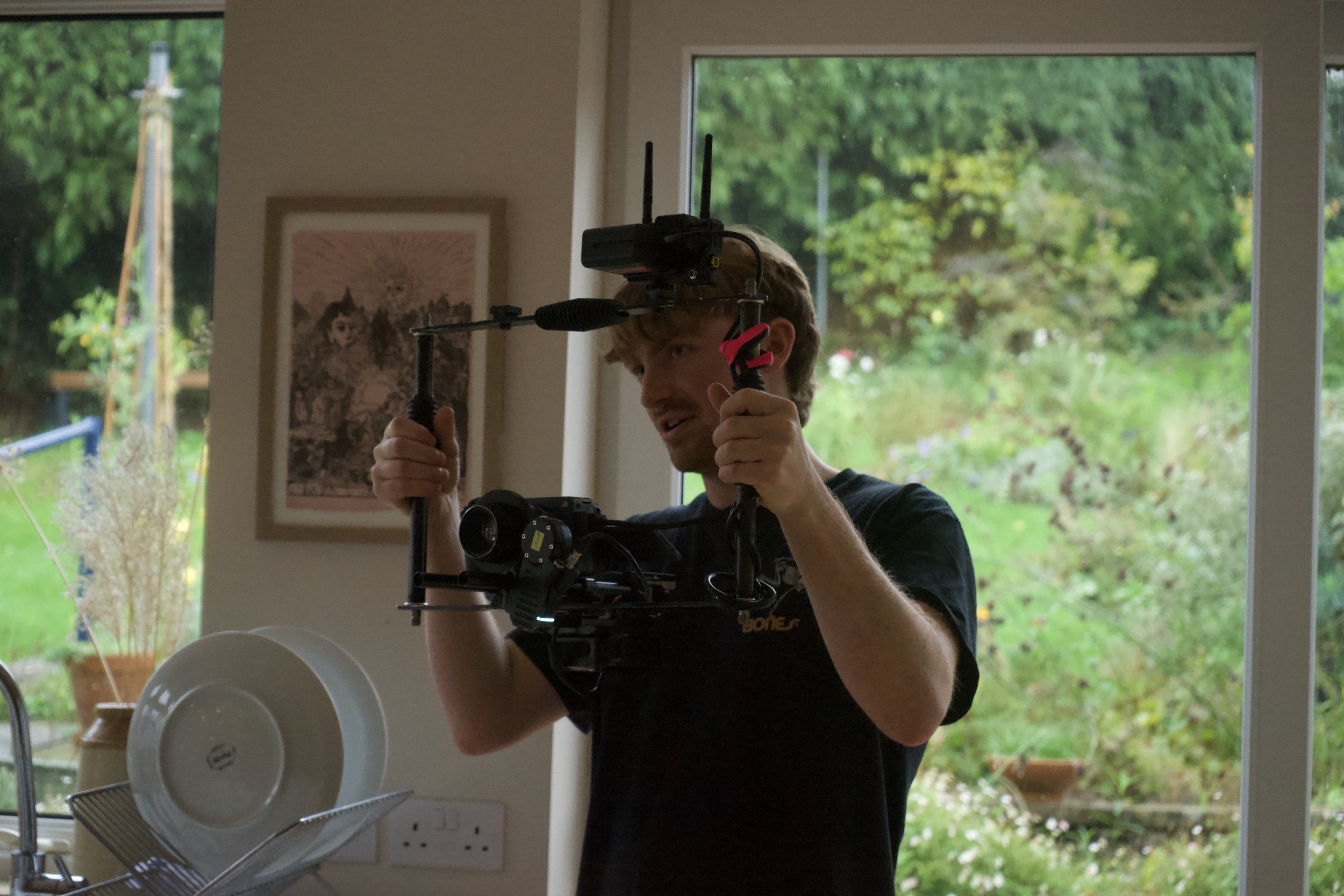 A man holding a camera crane with a mount for a camera and microphone, standing in a kitchen with large windows showing a green garden outside.