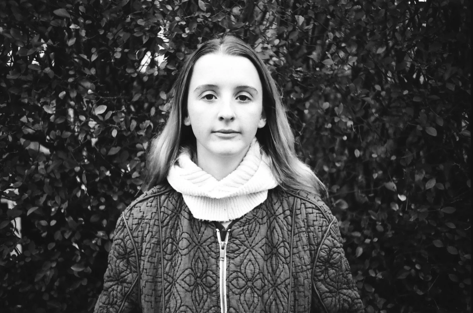 Black and white photo of a young girl with light-colored hair, wearing a quilted jacket with a zipper and a thick, knitted turtleneck sweater, standing in front of dense foliage.