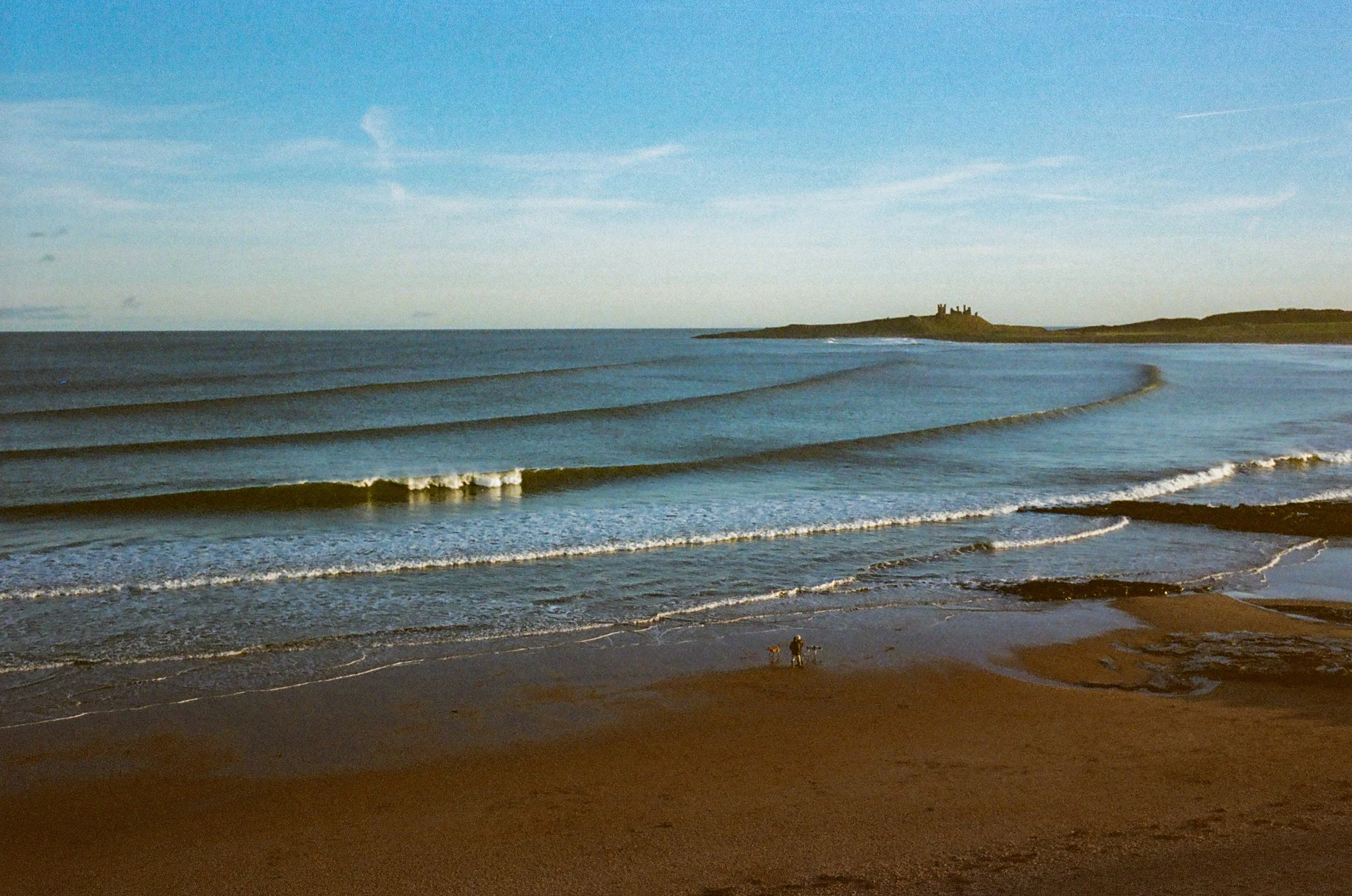 A sandy beach with gentle waves and a distant castle on a hill in the background under a blue sky.