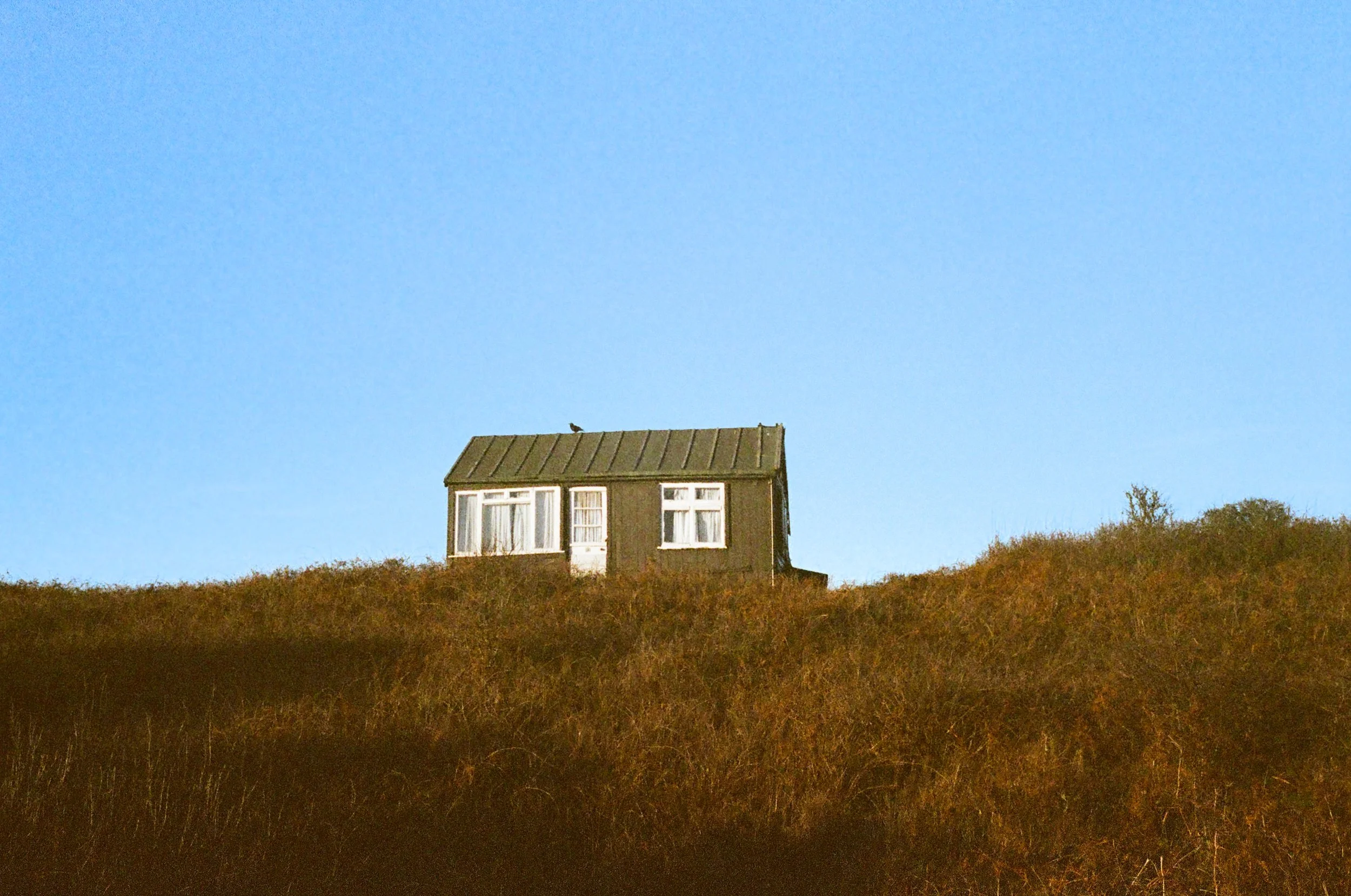 A small house on a hilltop with a grassy landscape, under a clear blue sky.