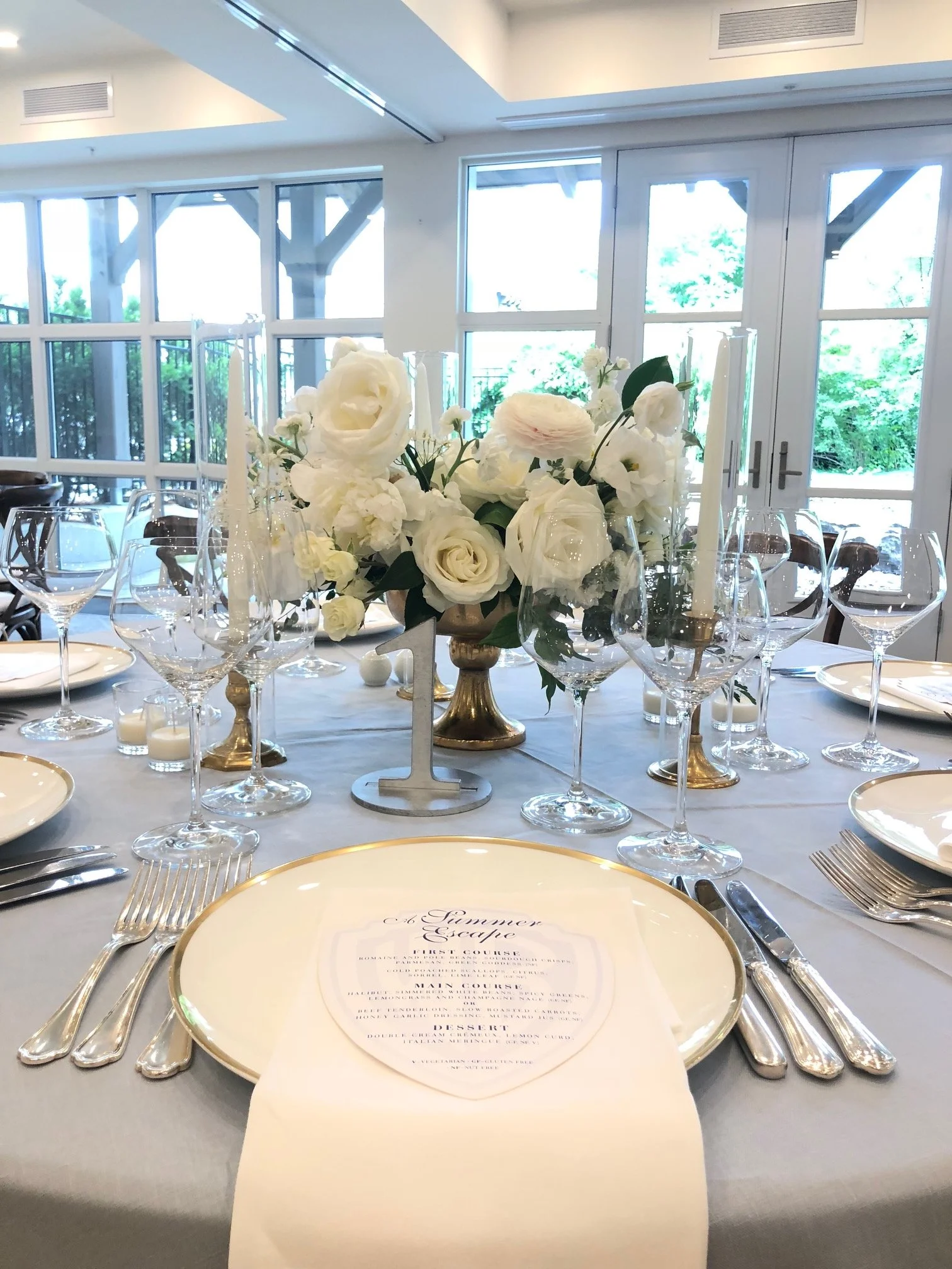 Elegant table setting with white flowers in a gold centerpiece, white plates, silverware, multiple wine glasses, and candles, set in a bright room with large windows and glass doors.