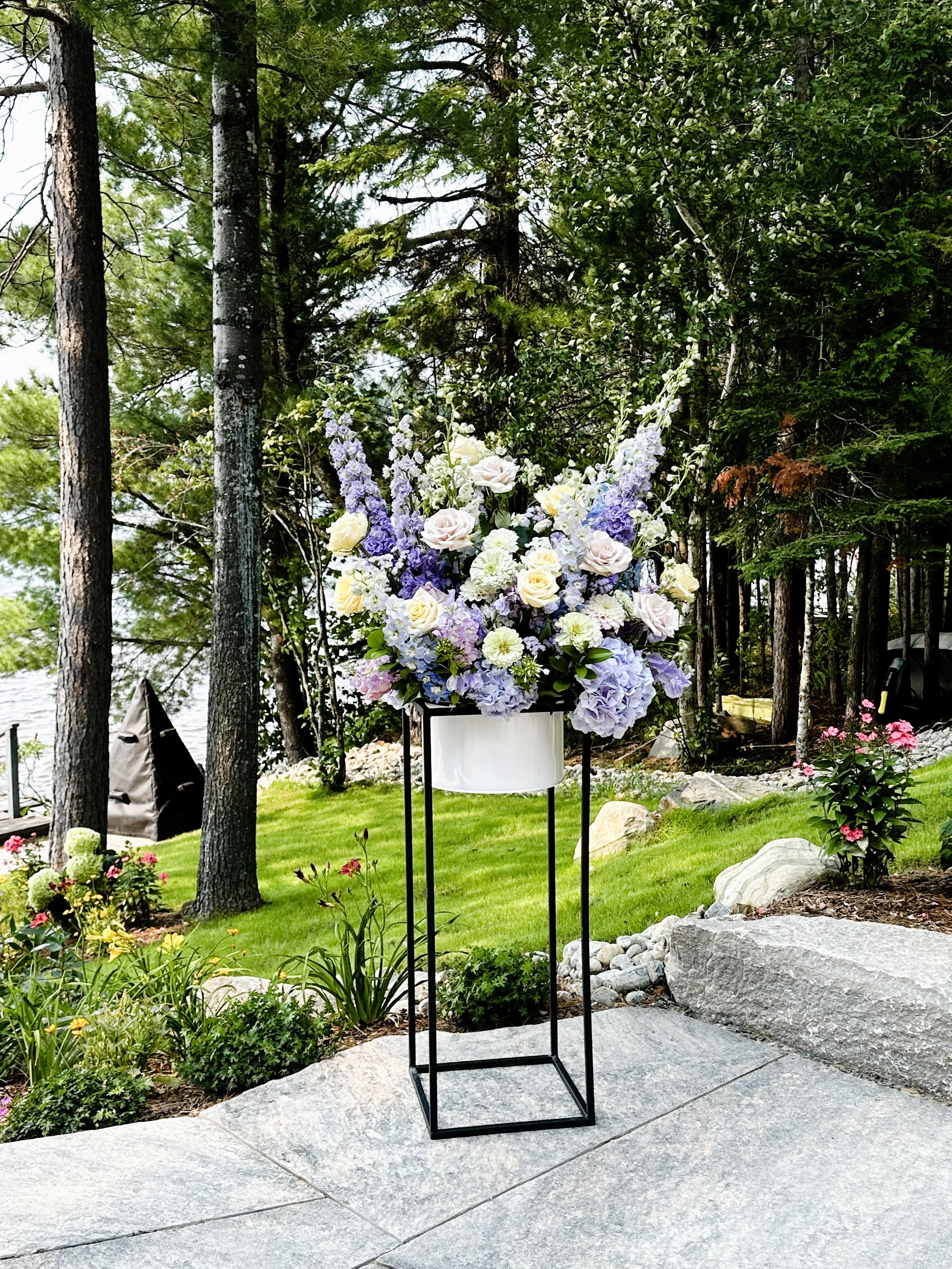 Large flower arrangement with white, purple, and pale pink flowers in a white vase on a black metal stand, outdoors near trees and a grassy area.