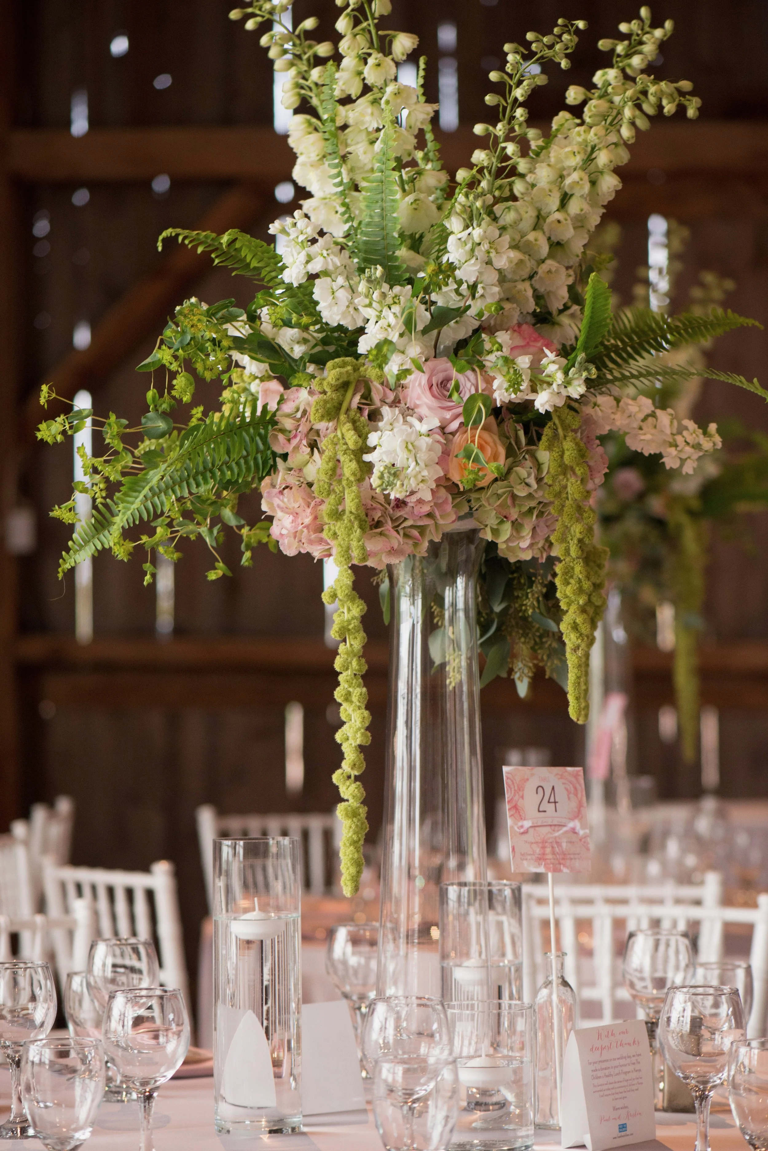 Tall floral centerpiece with white, pink, and green flowers and foliage on a table at a wedding reception, with candles and place cards.