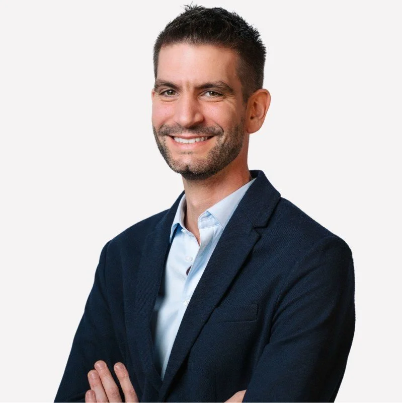 Homme d'affaires souriant, en costume noir et chemise blanche, avec les bras croisés contre un fond blanc.