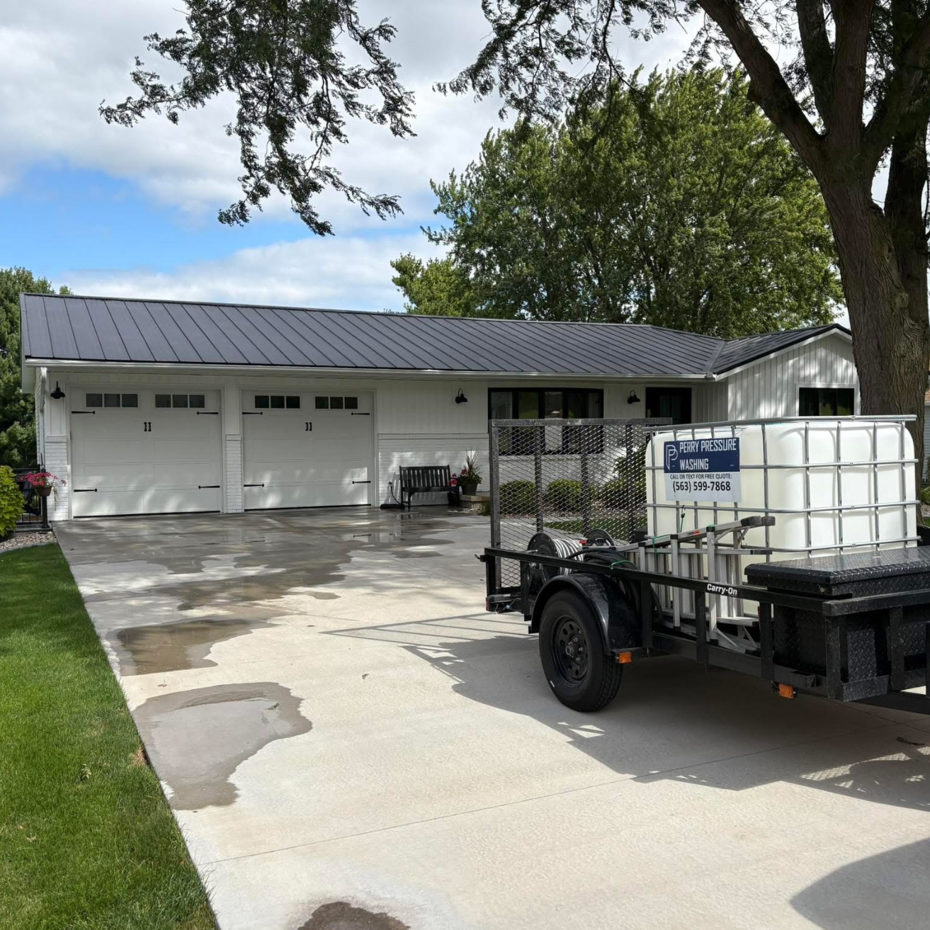 A white house with a two-car garage, a concrete driveway with wet spots, a green lawn, and a trailer with a water tank labeled 'Perry Pressure Washing' parked in front.