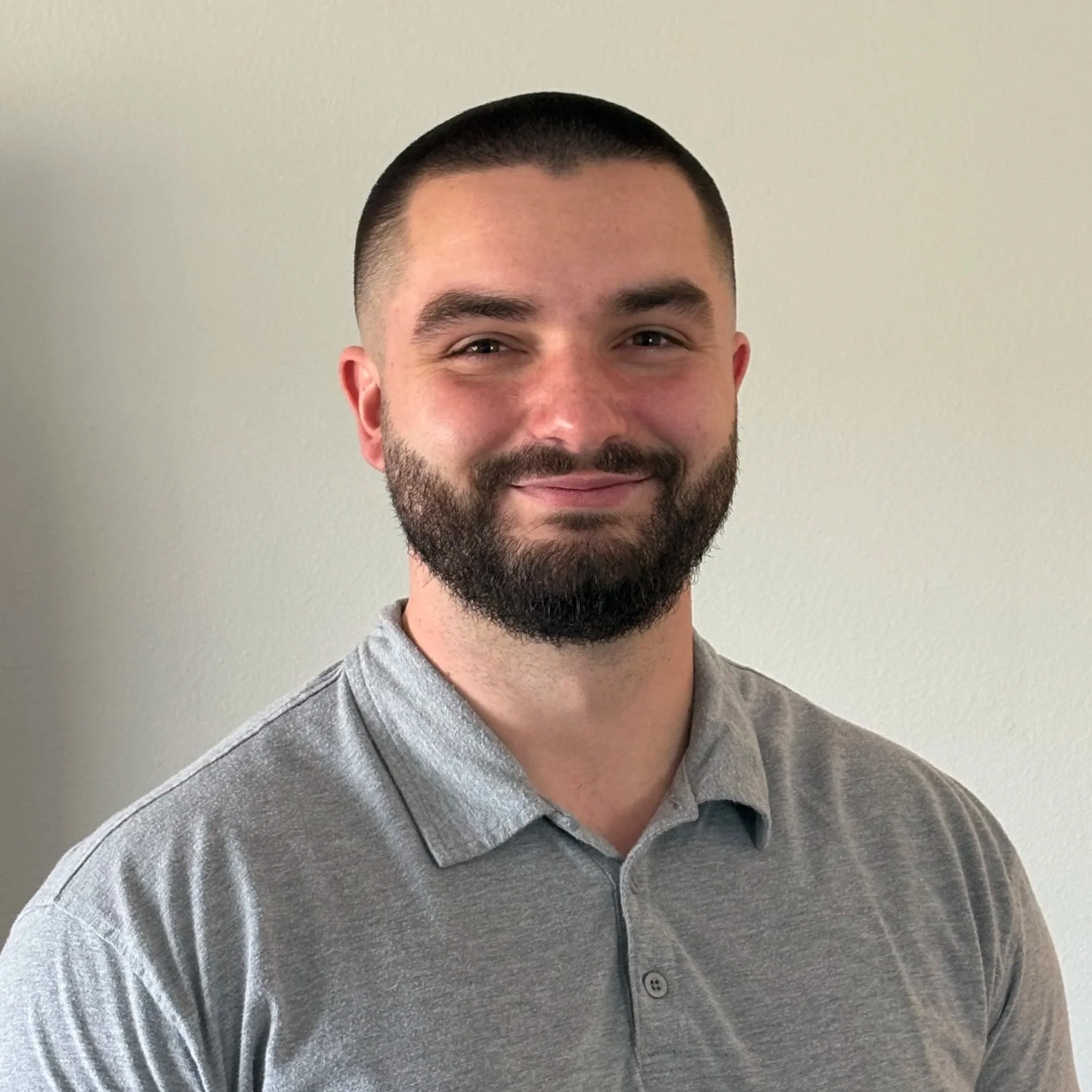 Portrait of a man with short dark hair, a full beard, wearing a gray collared shirt, smiling at the camera, against a plain light-colored background.