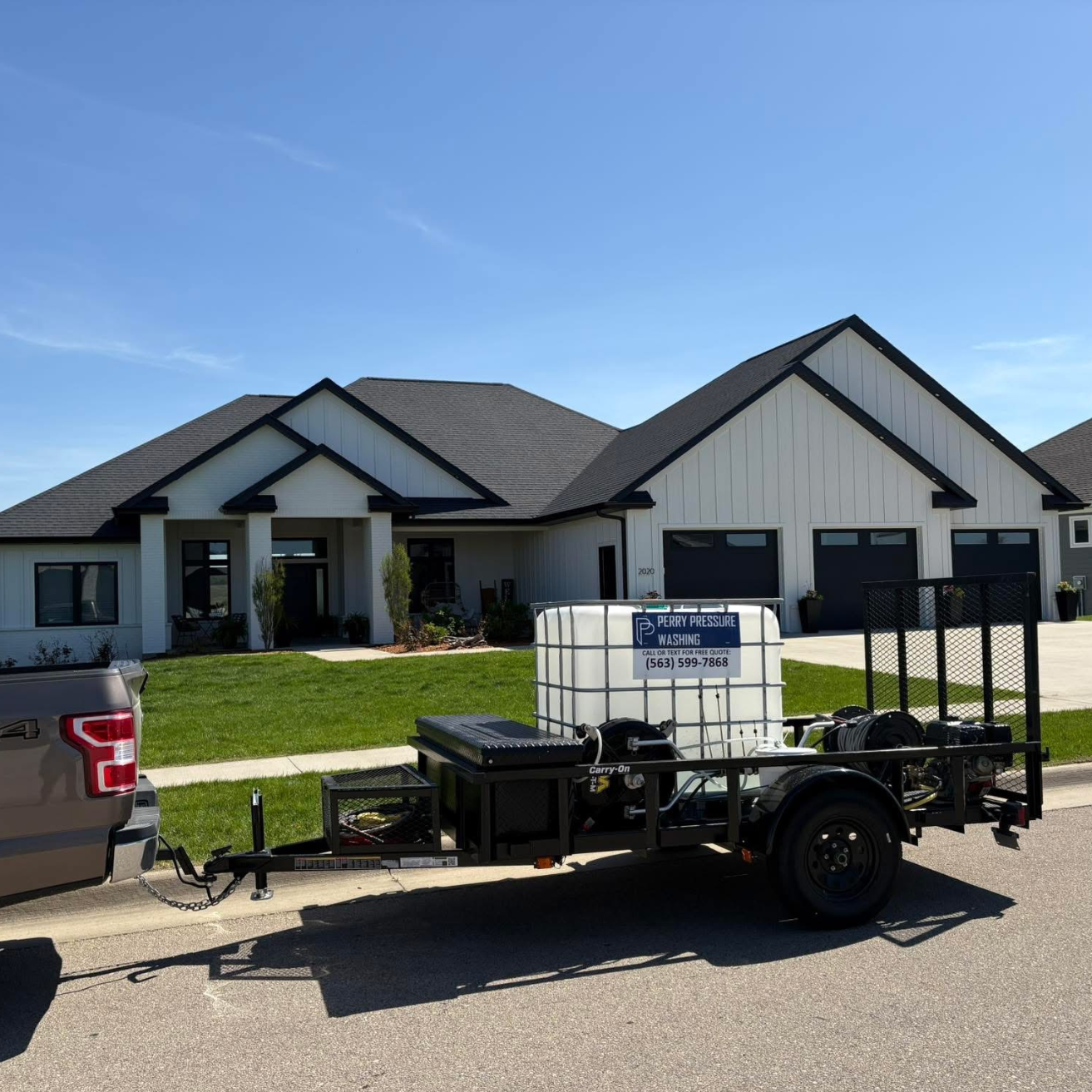 A house with a gray roof and white siding, with a front lawn, two garages, and a porch. A trailer with pressure washing equipment and a sign reading "Perry Pressure Washing" is parked in front of the house.