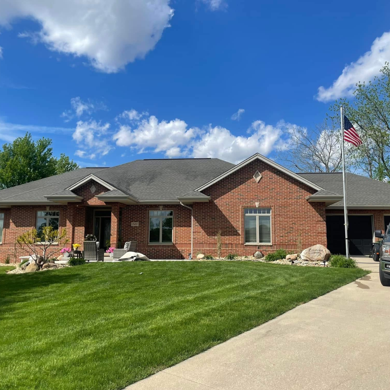 A single-story red brick house with a gray roof, a front porch with chairs, a lawn, a flagpole with an American flag, and a garage on a sunny day with blue sky and clouds.