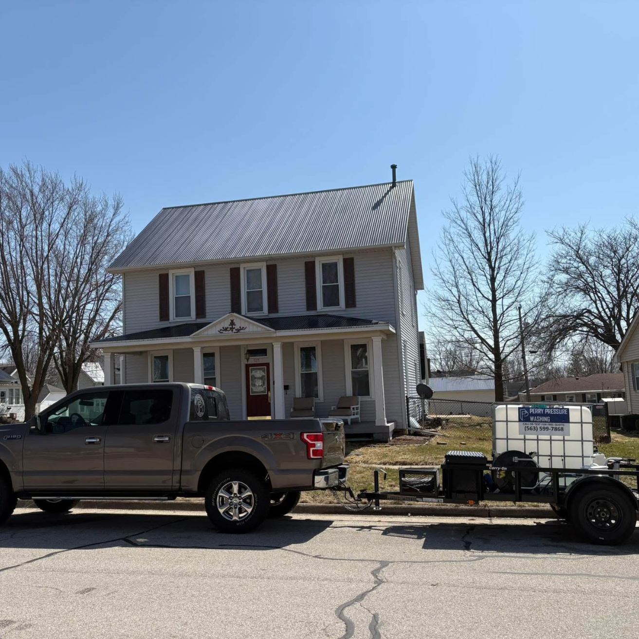 A gray pickup truck parked on a residential street in front of a white two-story house with brown shutters and a front porch. Attached to the truck is a trailer with equipment and a sign that reads "Perry Pressure Washing" with phone number 563-599-7868.