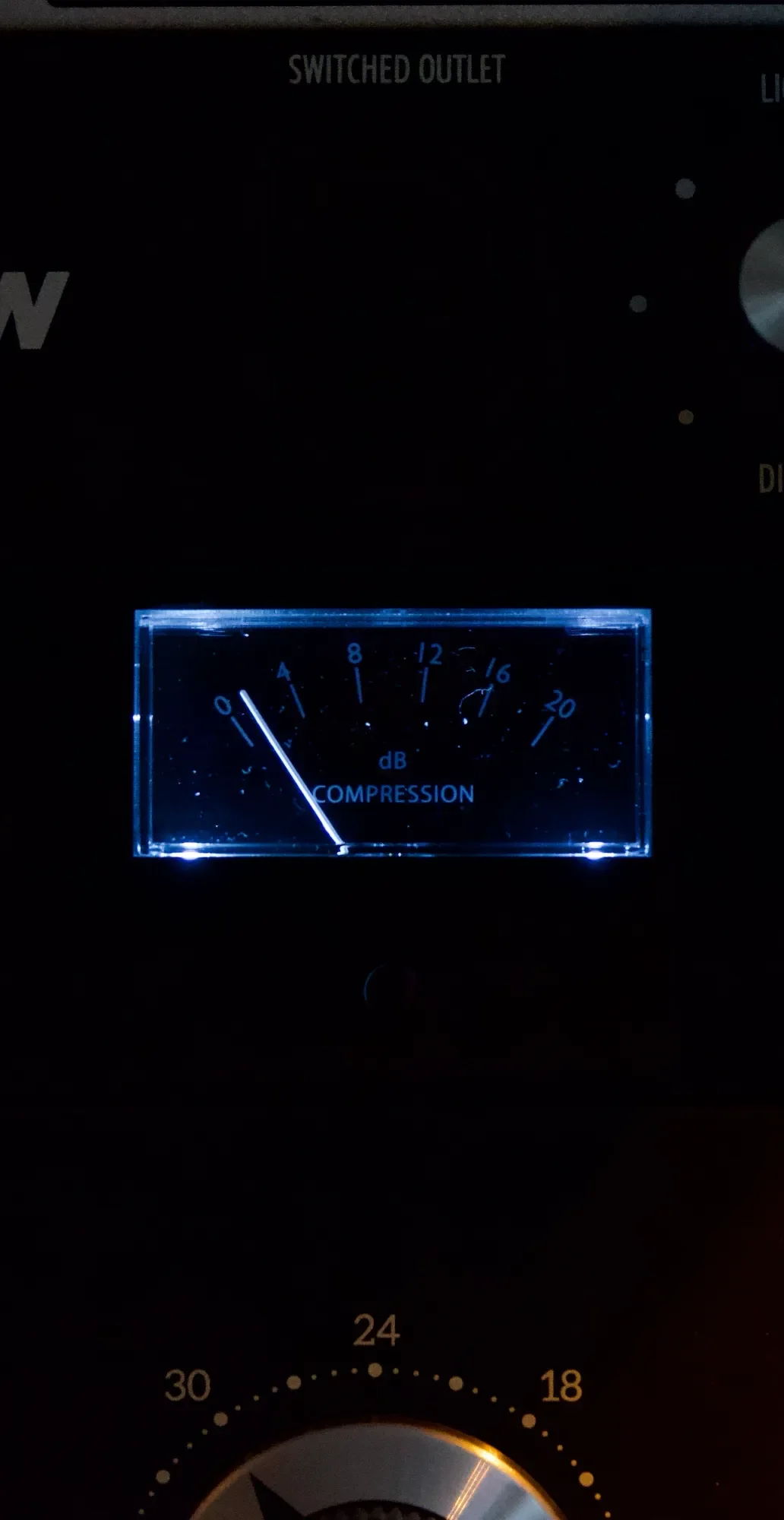 A close-up of an audio equipment panel displaying a blue backlit compression level gauge with a needle pointing to 1. The gauge shows decibel levels from 0 to 20, with the labels 'dB' and 'COMPRESSION.' Above the gauge, the panel reads 'SWITCHED OUTLET.'