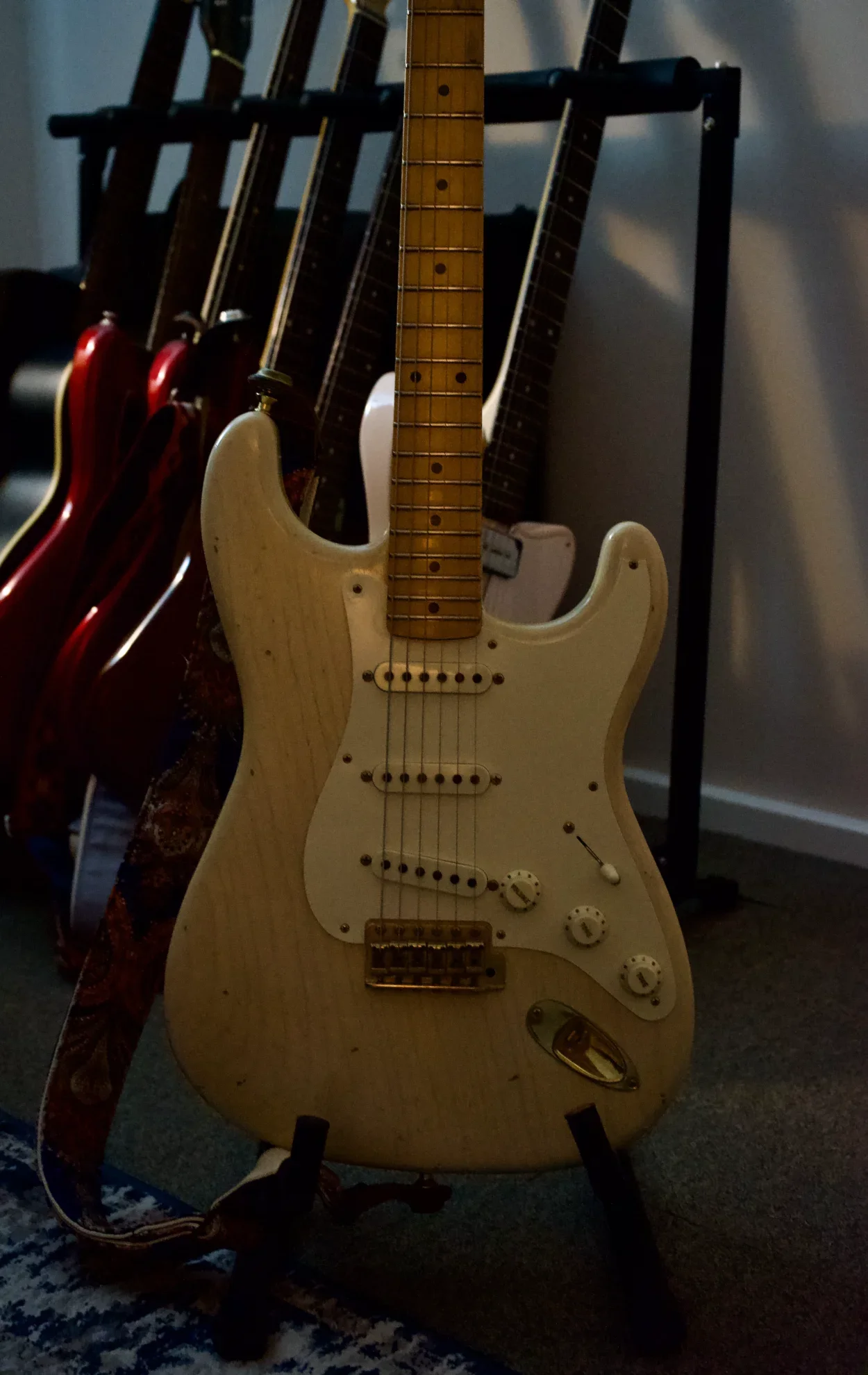 Cream-colored electric guitar on a stand, with other guitars hanging on a rack in the background.