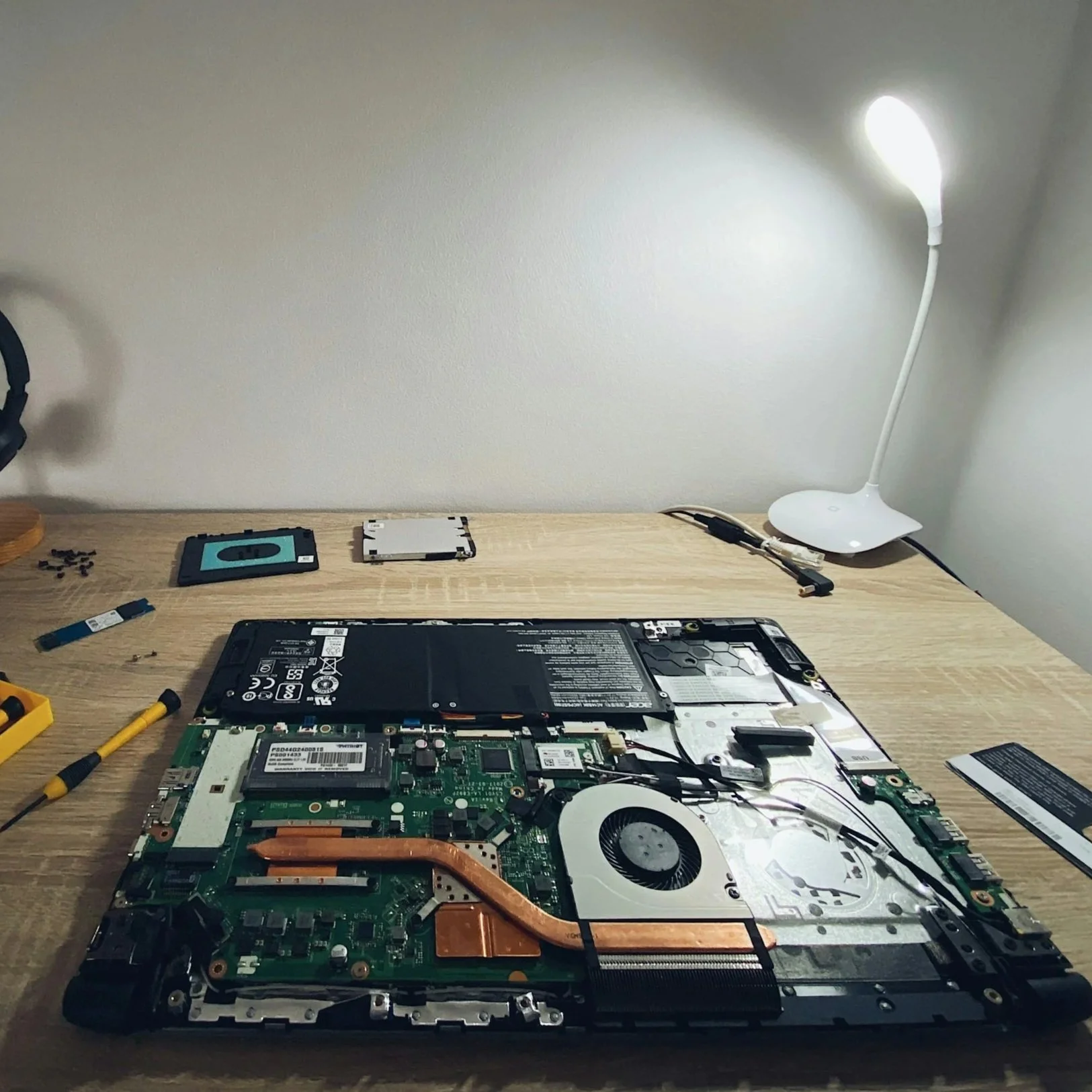 Disassembled laptop with internal components exposed on a wooden desk, tools around, and a white lamp illuminating the workspace.