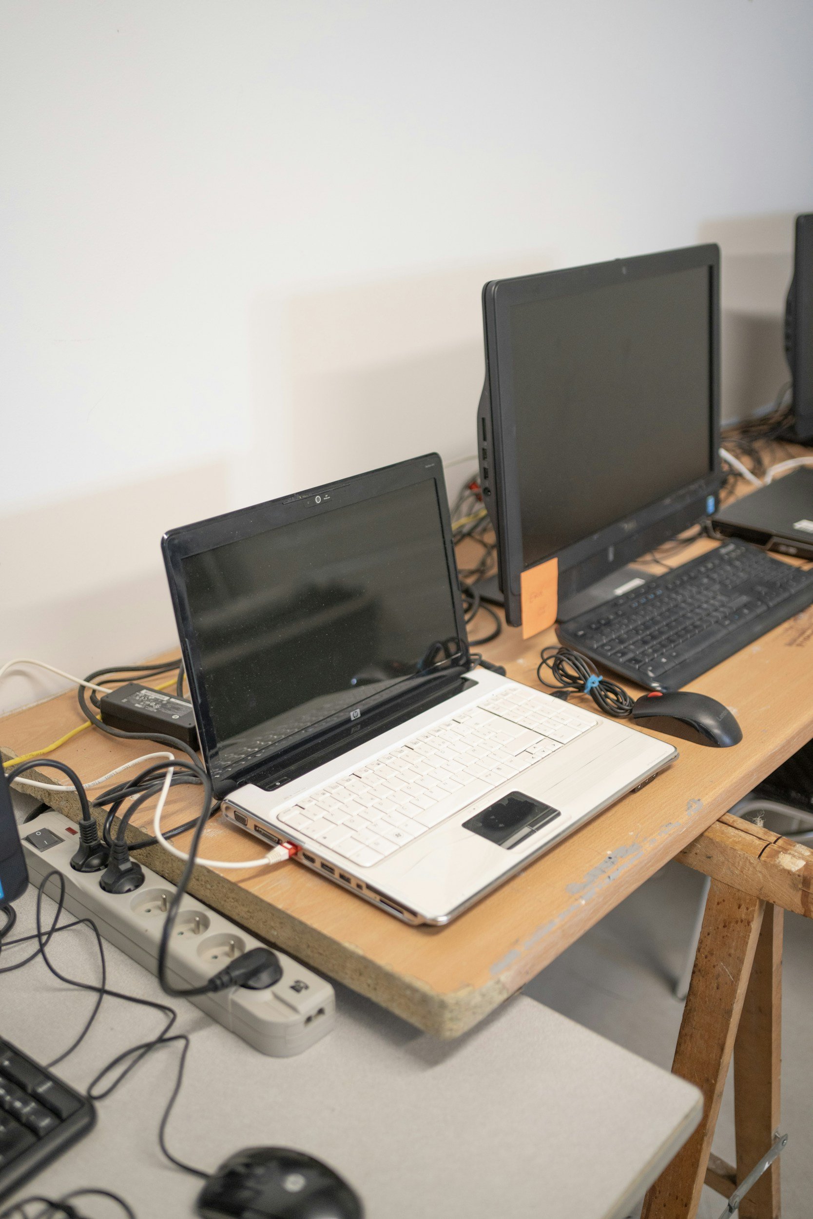 A wooden desk with multiple computers, a keyboard, mouse, power strip, and various cables in an office setting.