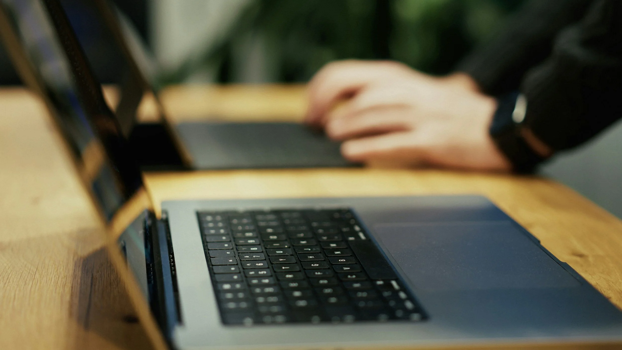 Close-up of a person's hand using a laptop with a wooden surface, with blurry background.