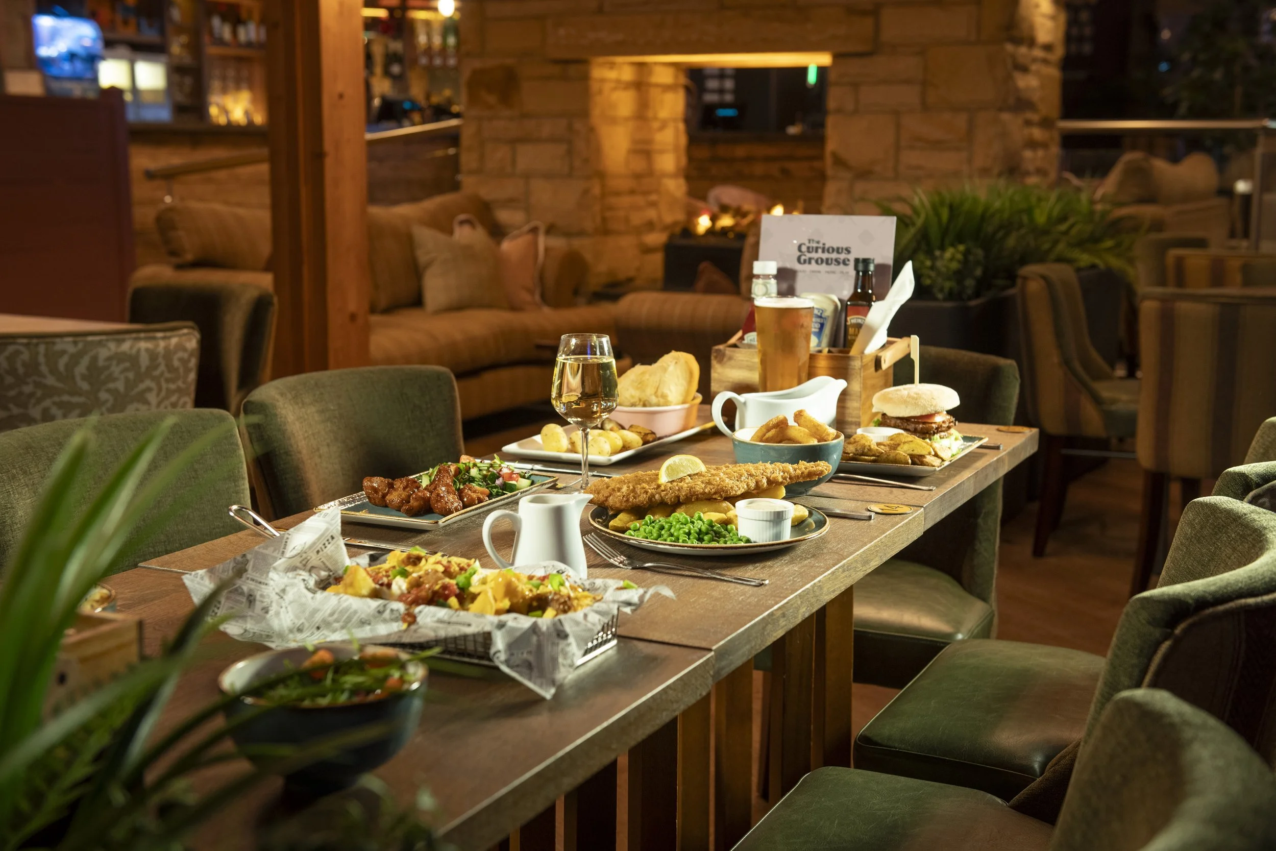 A table full of plates of food: fish and chips with garden peas, cauliflower bites, loaded chips, onion rings, veggie burger and a stake pie. There is a glass of white wine and a pint of beer in between the dishes.