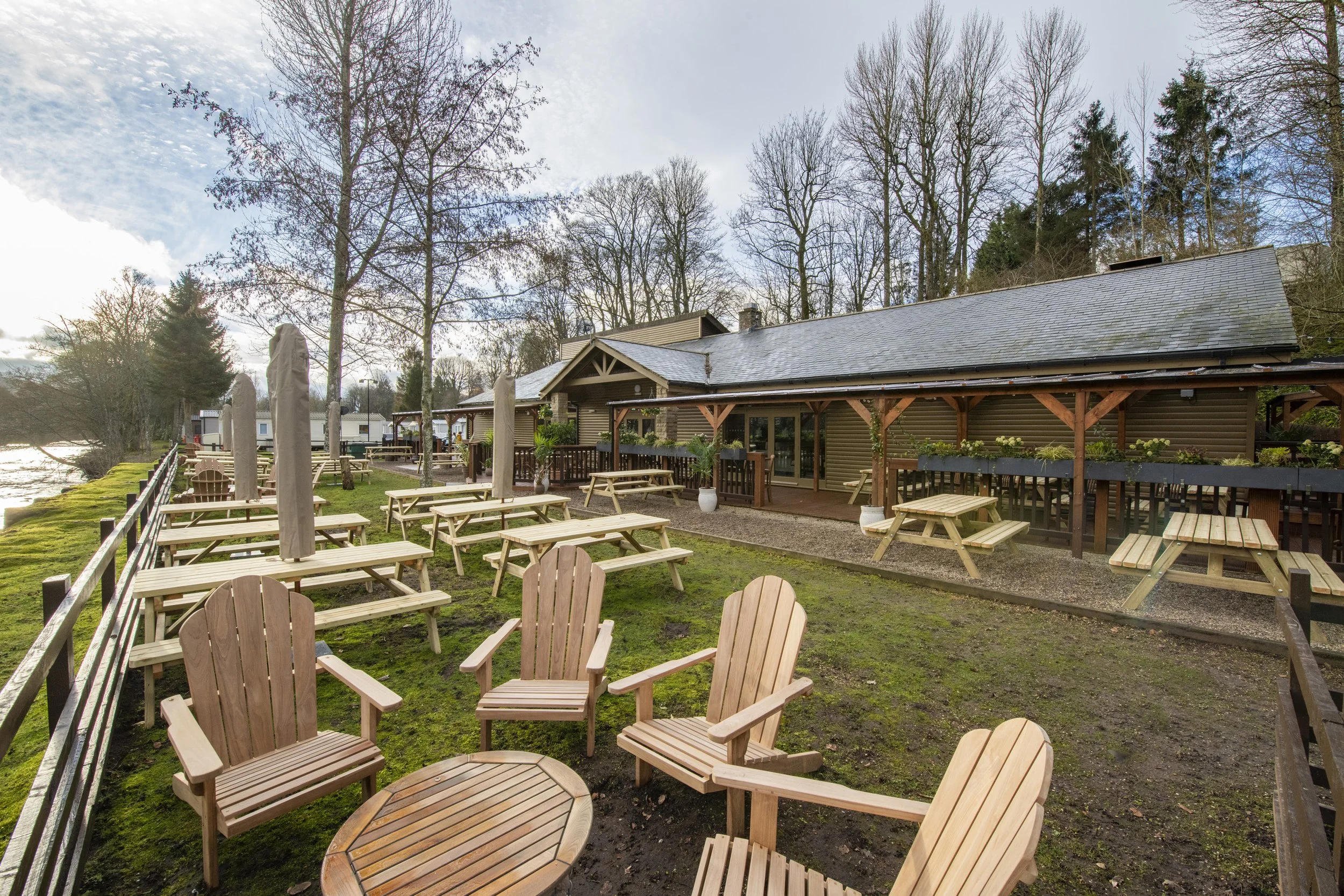 A table set for breakfast with two plates of eggs, bacon, sausage, pancakes, mushrooms, baked beans, toast, and drinks including coffee, orange juice, and a latte. Behind the table is a scenic riverside view with a wooden building, outdoor seating, picnic tables, trees, and a river with rocks.