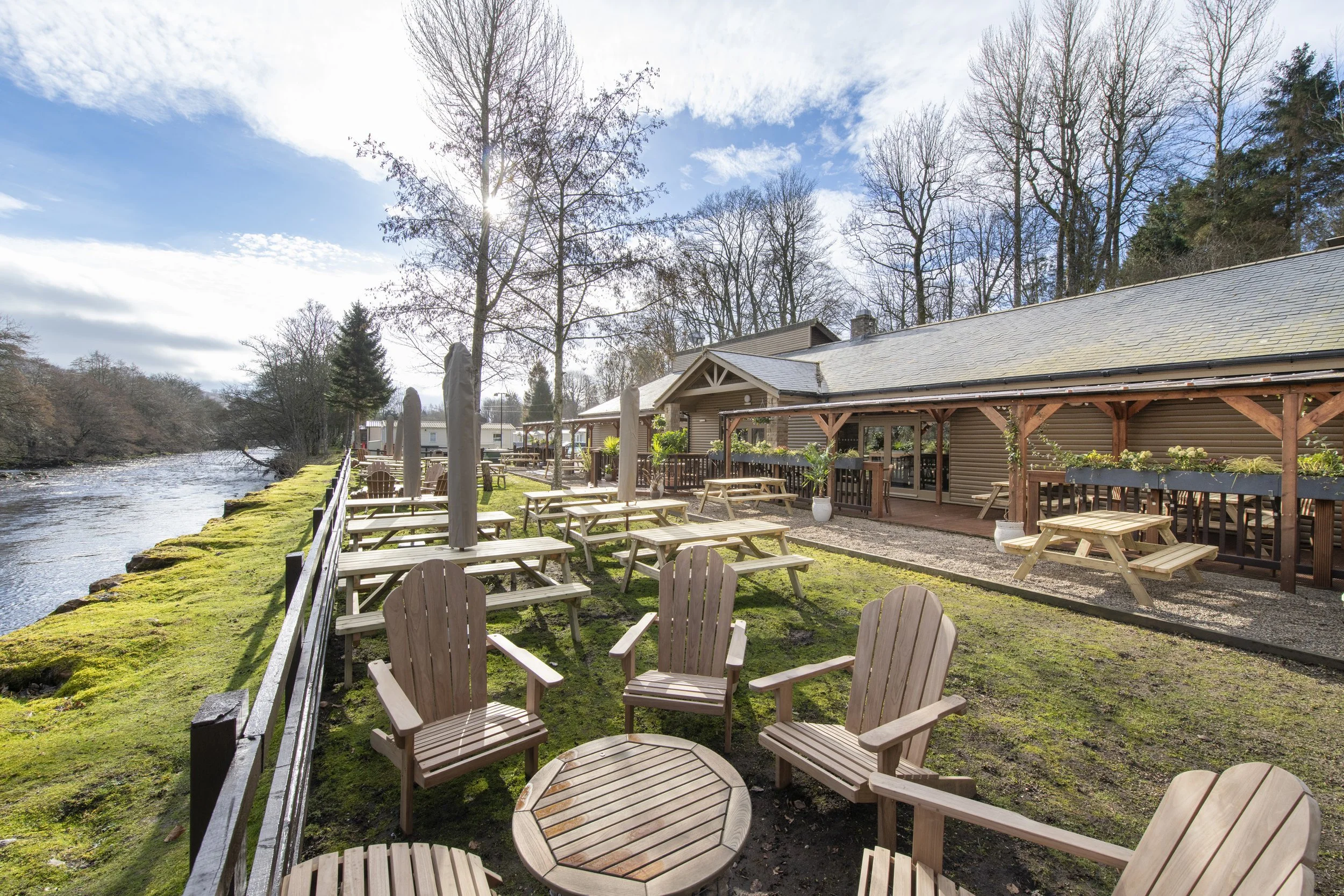 A wooden building with outdoor seating near a river, surrounded by green trees, during the evening with lights lit inside.