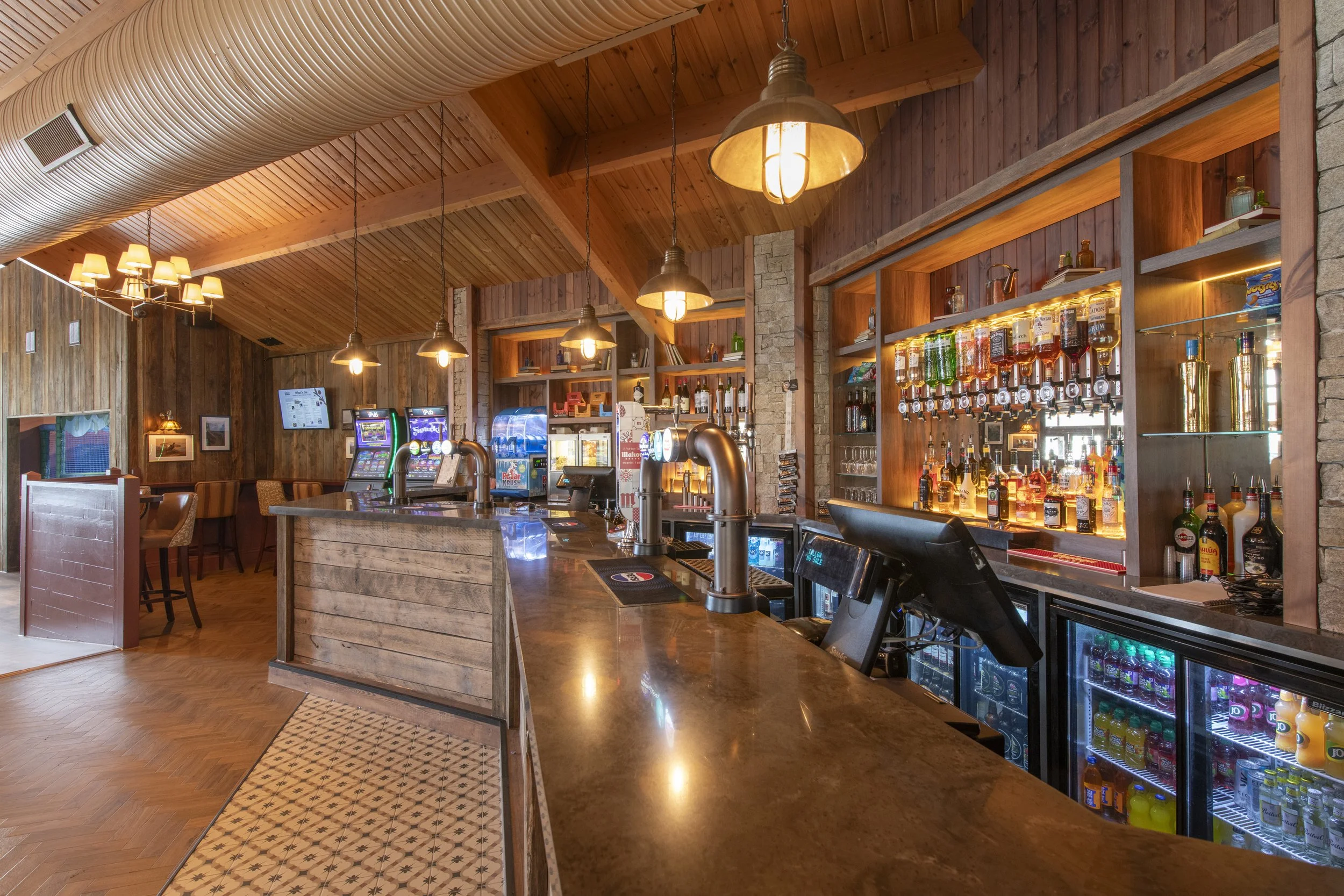 A full bar, featuring wood panels on walls and brick features. The bar is stocked with glass bottles of liquor, bottles of juice and glassware.