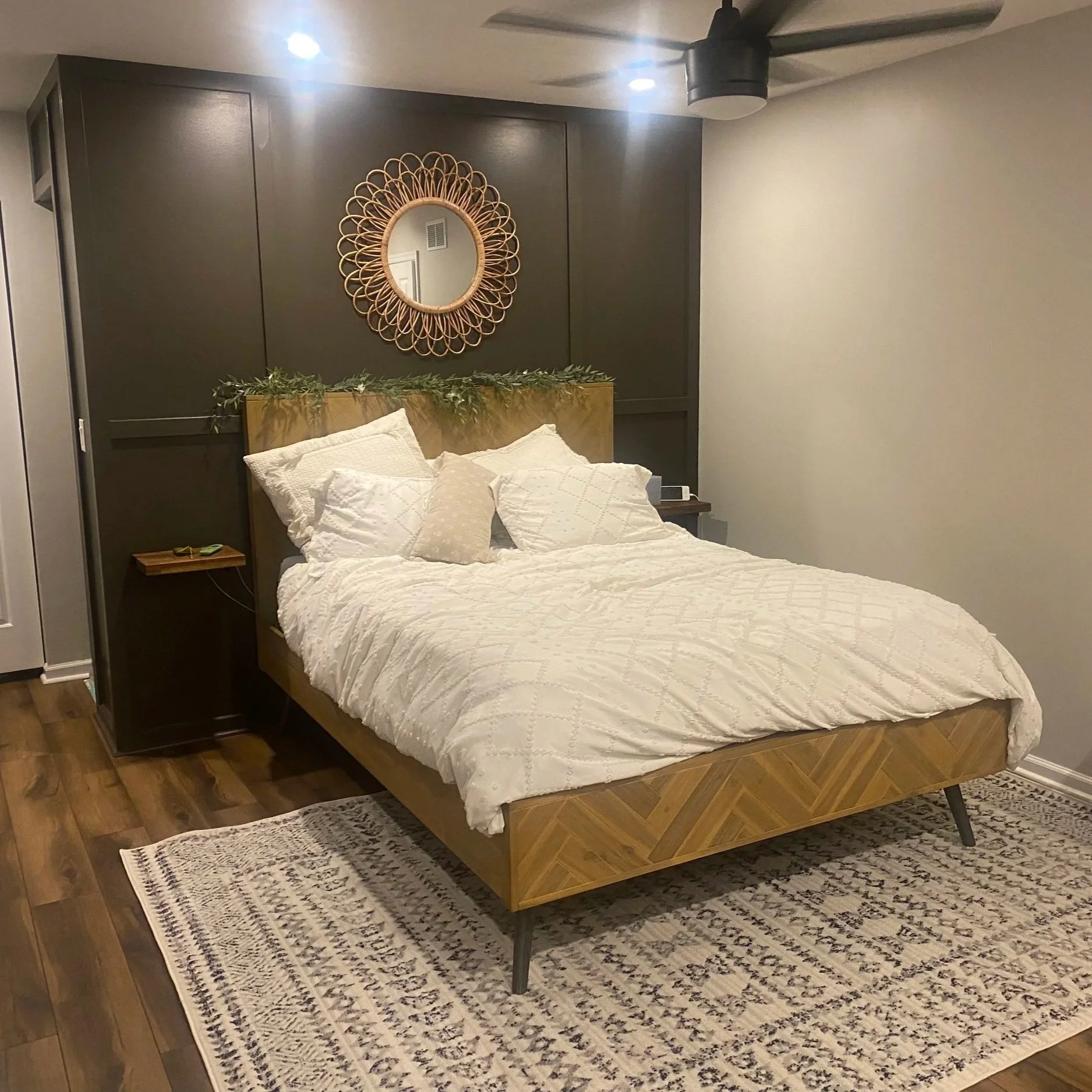 A bedroom with a bed, white bedding, and pillows, on a patterned rug; dark paneled wall with a round decorative mirror and greenery above the headboard, and a ceiling fan above.