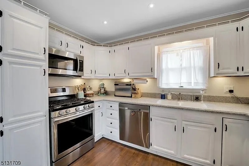 Kitchen with white cabinets, stainless steel appliances including a microwave, stove, and dishwasher, granite countertops, a window above the sink with a curtain, and hardwood flooring.
