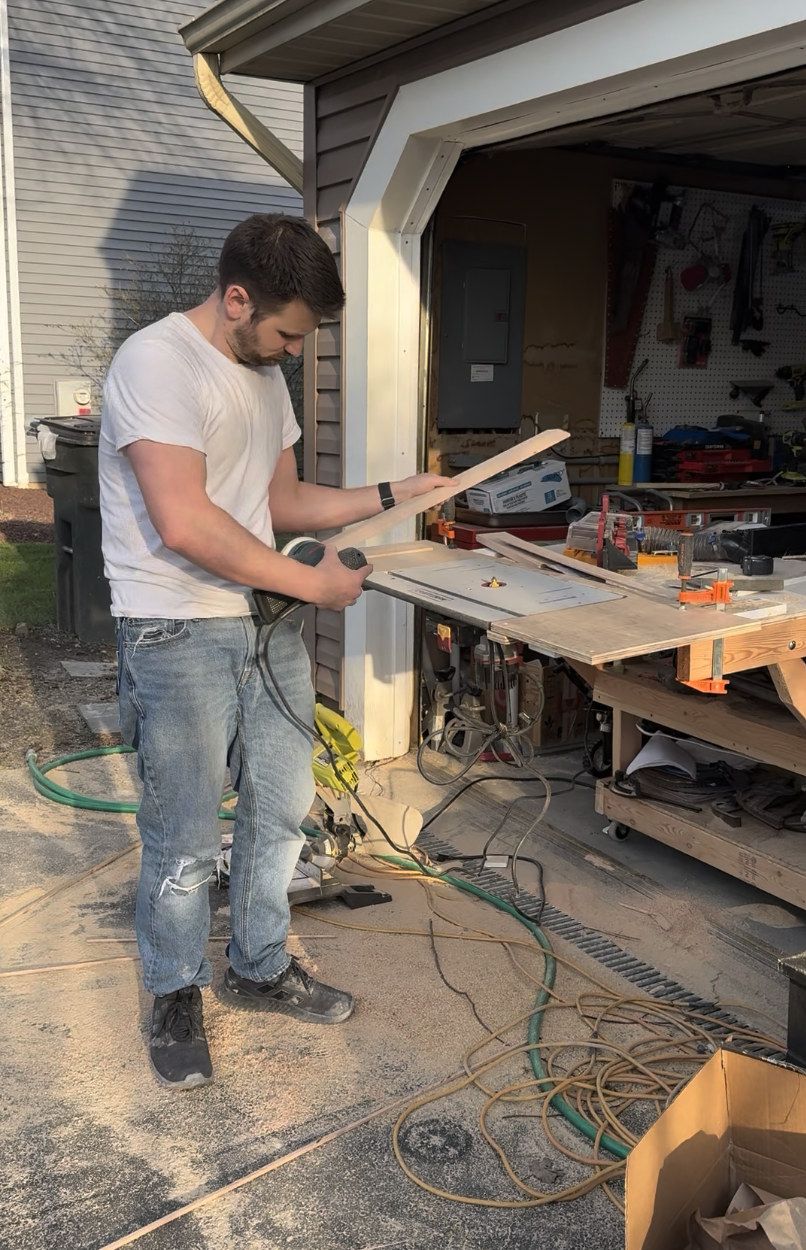 A man working on a woodworking project outside a garage. He is holding a power saw and a piece of wood, surrounded by tool clutter and extension cords.