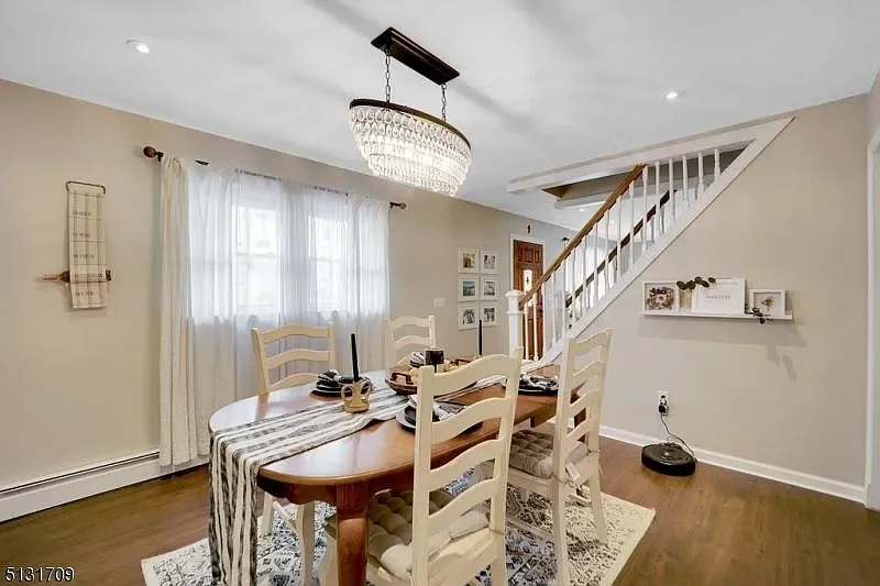 Dining room with a wooden table and six chairs, chandelier lighting, a window with curtains, staircase with white railing, wall shelves with decor, and a robotic vacuum on hardwood floor.